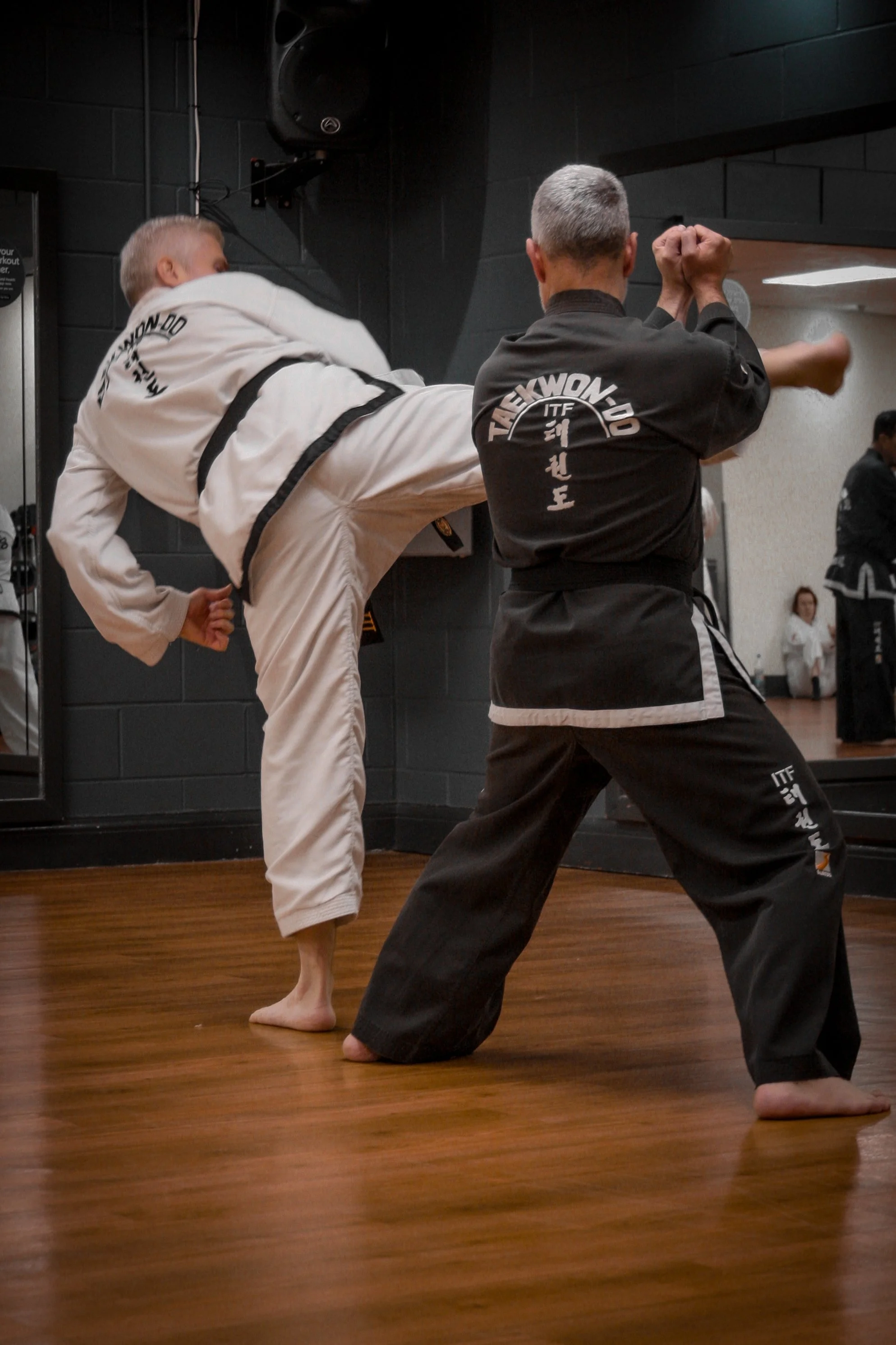 Two martial artists practicing Taekwondo in a training room. One is delivering a high kick, and the other is defending. Both are wearing black and white uniforms respectively with Taekwondo patches.