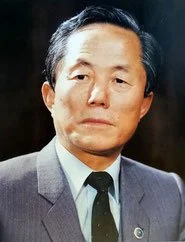 A middle-aged man with black hair wearing a gray suit and tie, looking serious.
