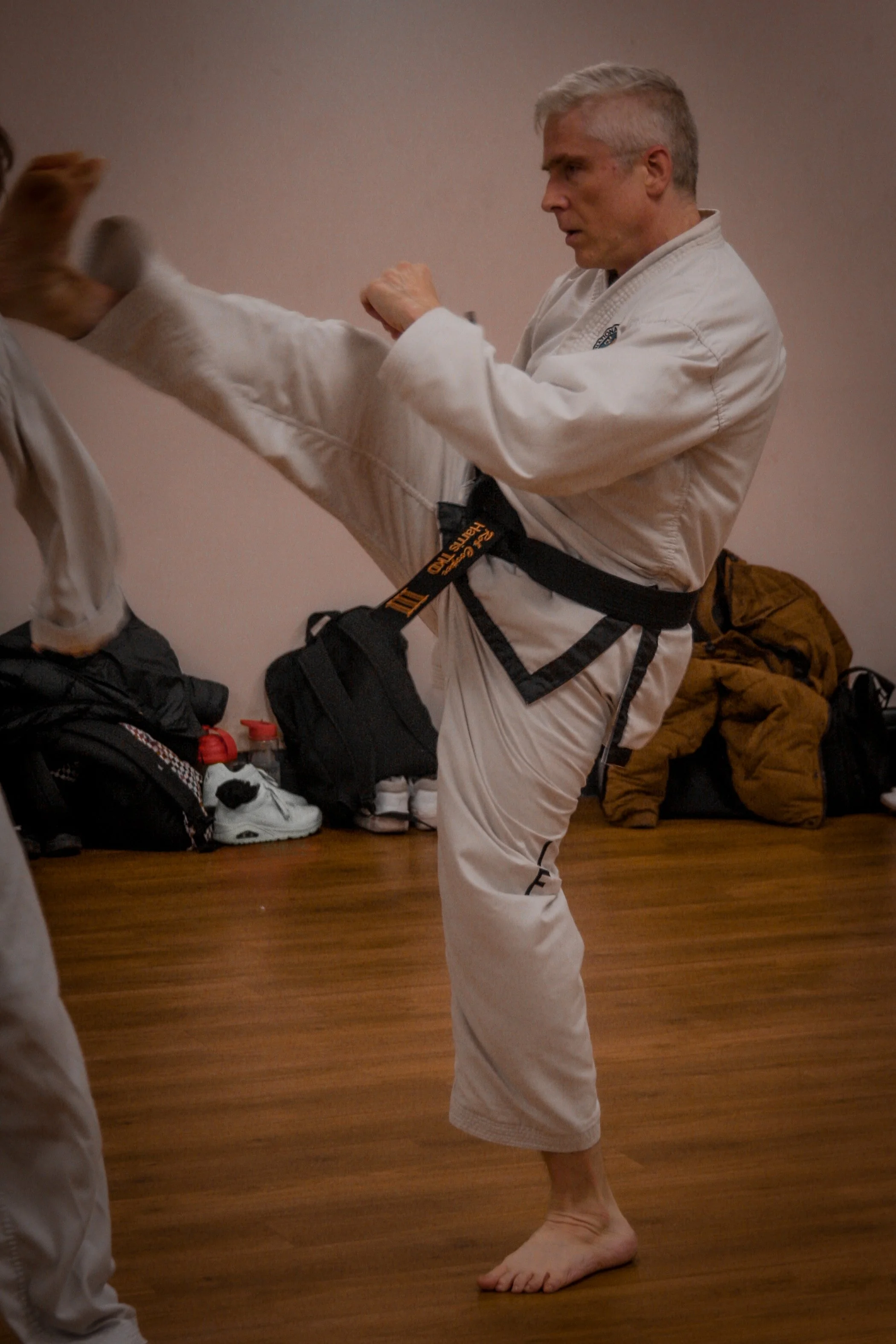 Martial artist practicing a high kick in a dojo, wearing a white gi and black belt, with training gear and bags in the background.