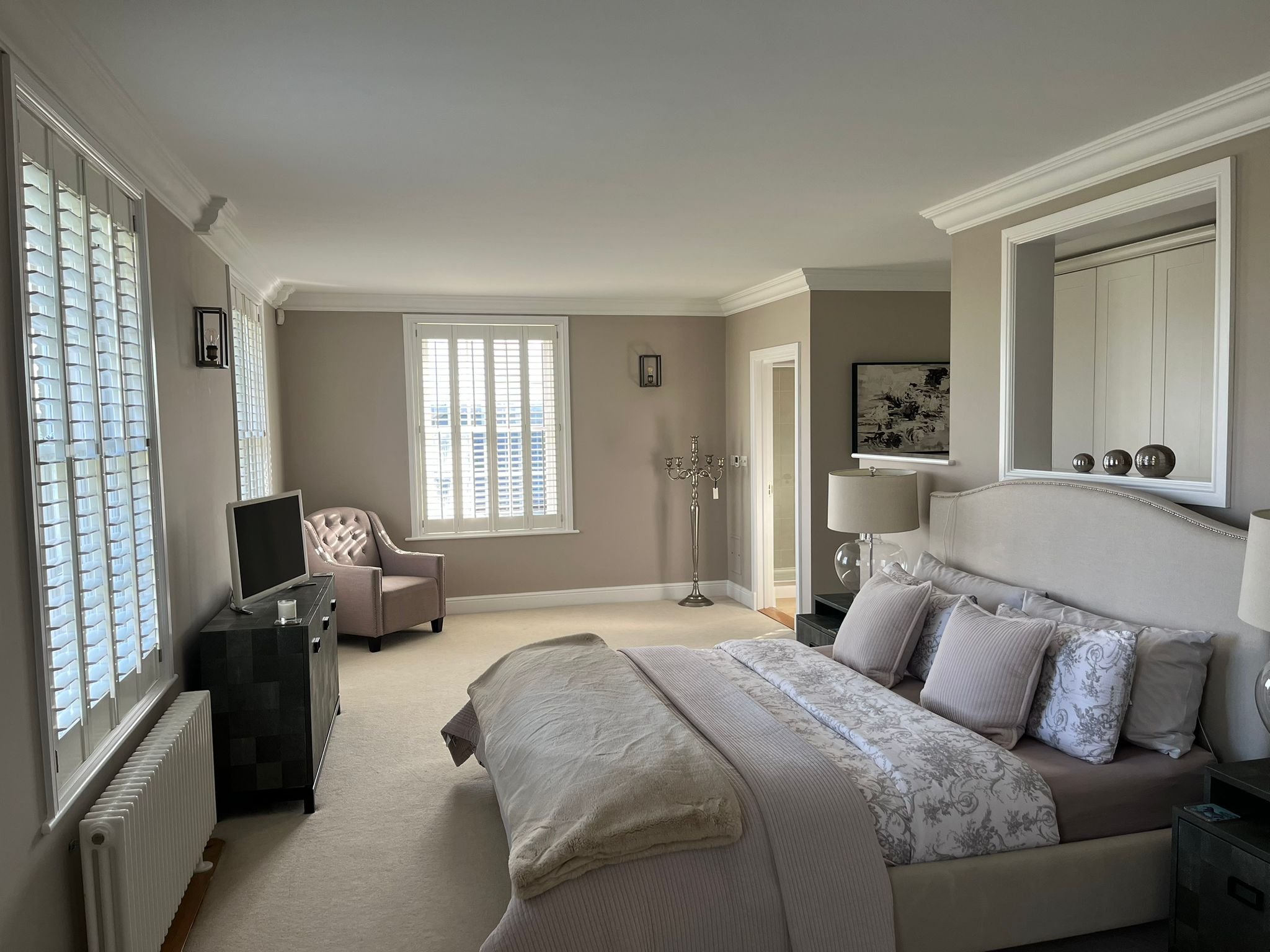 Bright bedroom with a large bed, white headboard, and multiple pillows. Nightstands with lamps, a window with white shutters, a small TV on a stand, a pink accent chair, and a beige carpeted floor.
