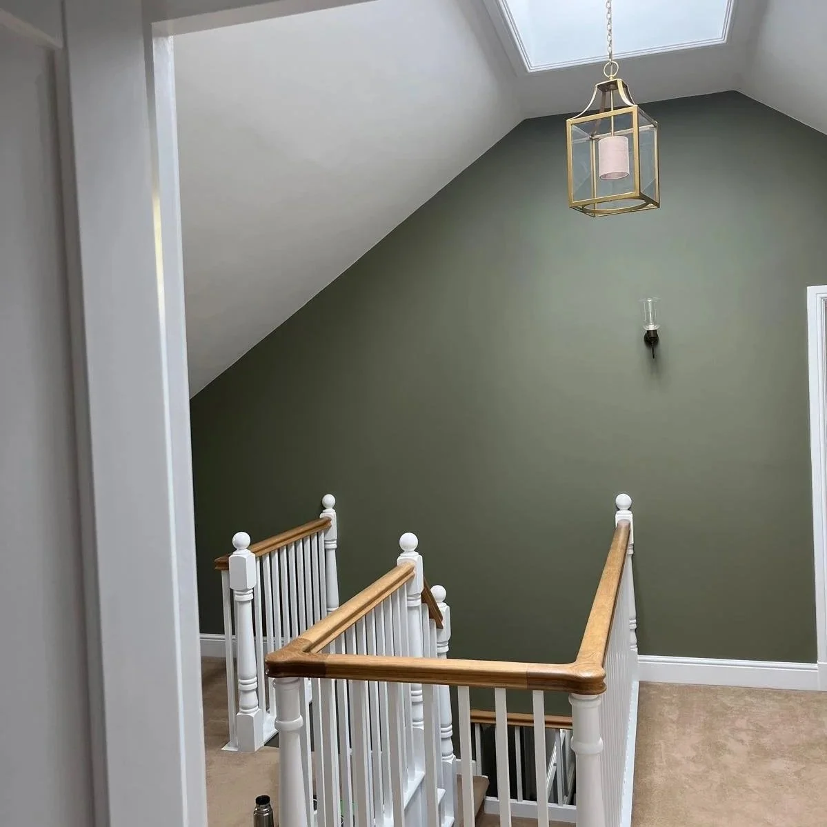 Interior view of a house staircase landing with a green wall, white railing with wooden handrails, a hanging light fixture, a wall sconce, and a skylight.
