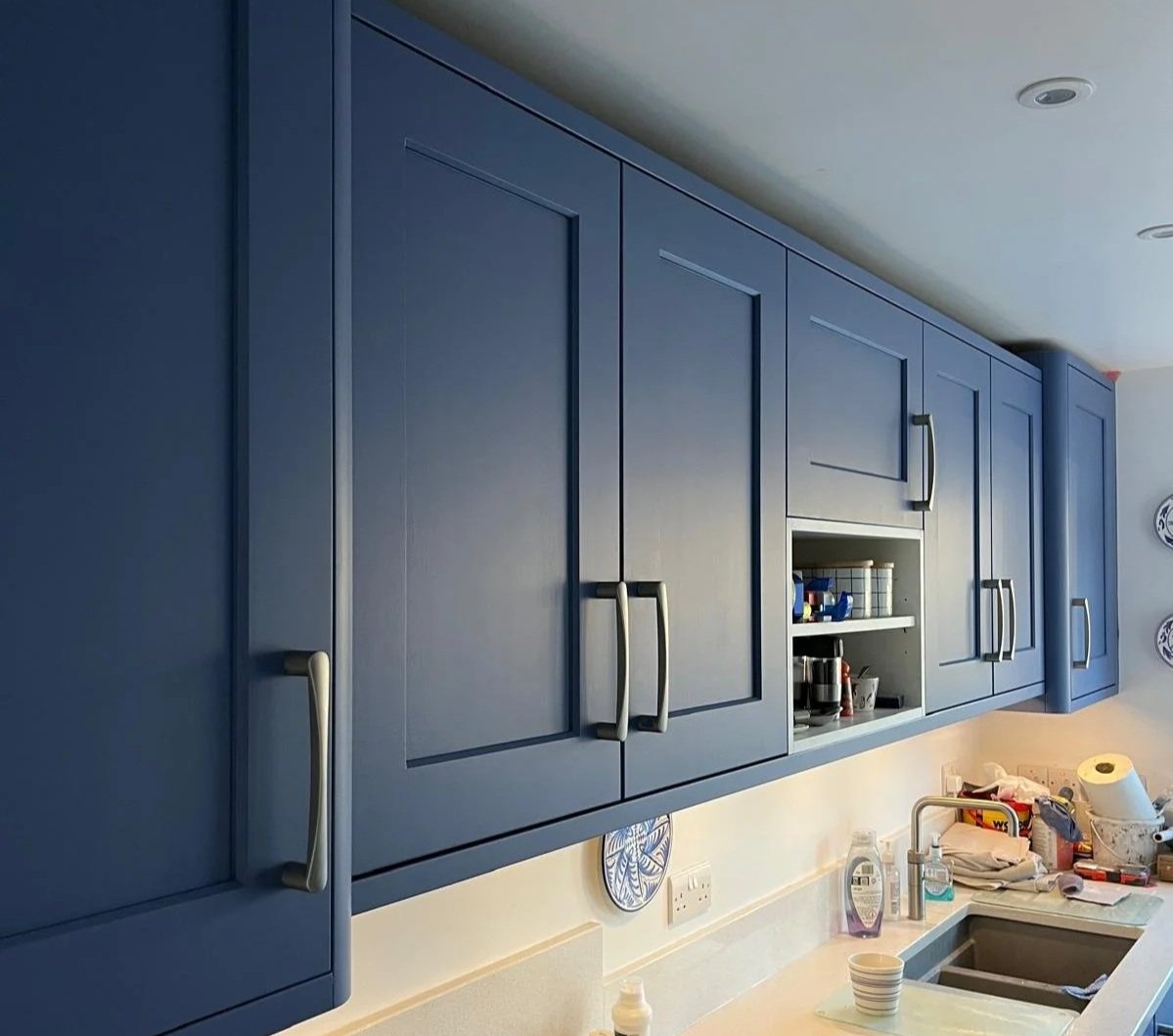 Blue kitchen cabinets above a beige countertop with scattered items including a paper towel roll, cups, and cleaning supplies.