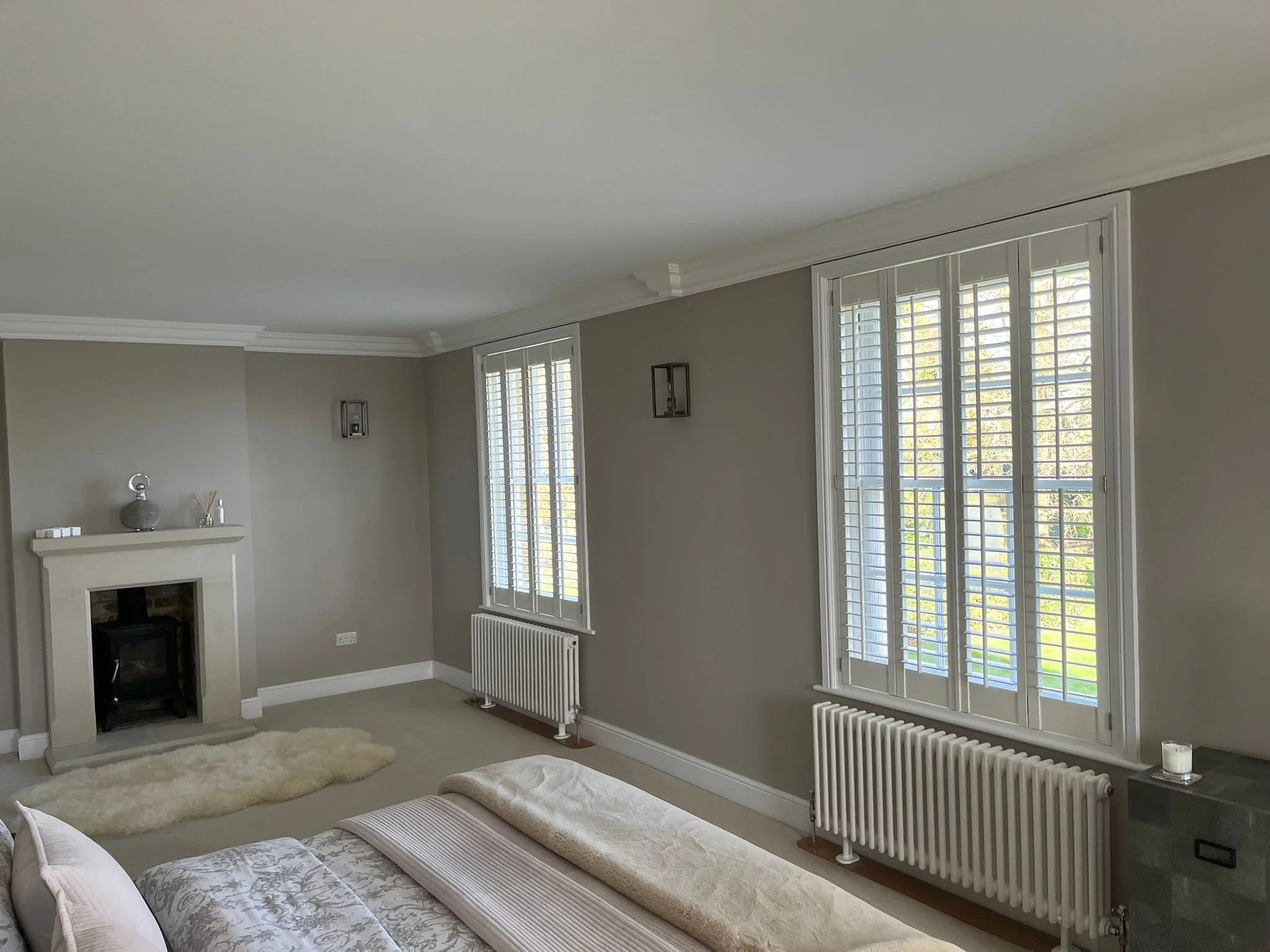 A living room with beige walls, white crown molding, and large windows with white shutters, a white fireplace mantle with decorative items, a cream-colored shaggy rug, part of a beige and white bed, and a radiator under the windows.
