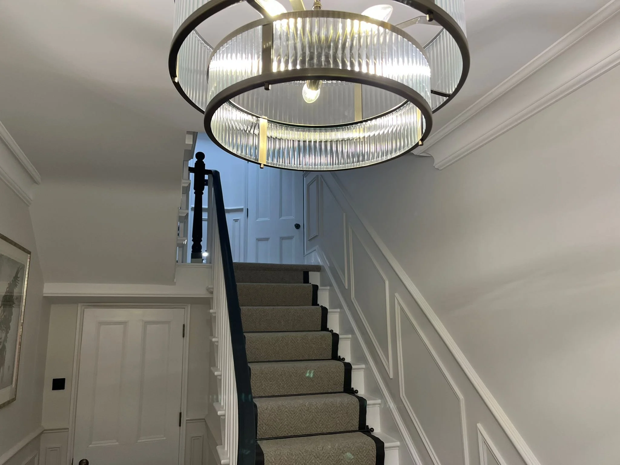 Interior view of a staircase with white walls, beige carpeted stairs, black handrail, and a modern circular chandelier hanging from the ceiling.