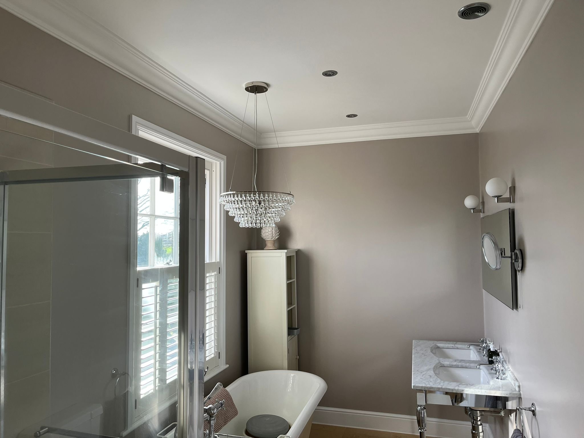 Bathroom with a clawfoot bathtub, a glass shower enclosure, a double sink marble vanity, a white cabinet, and a chandelier. Light gray walls and crown molding.