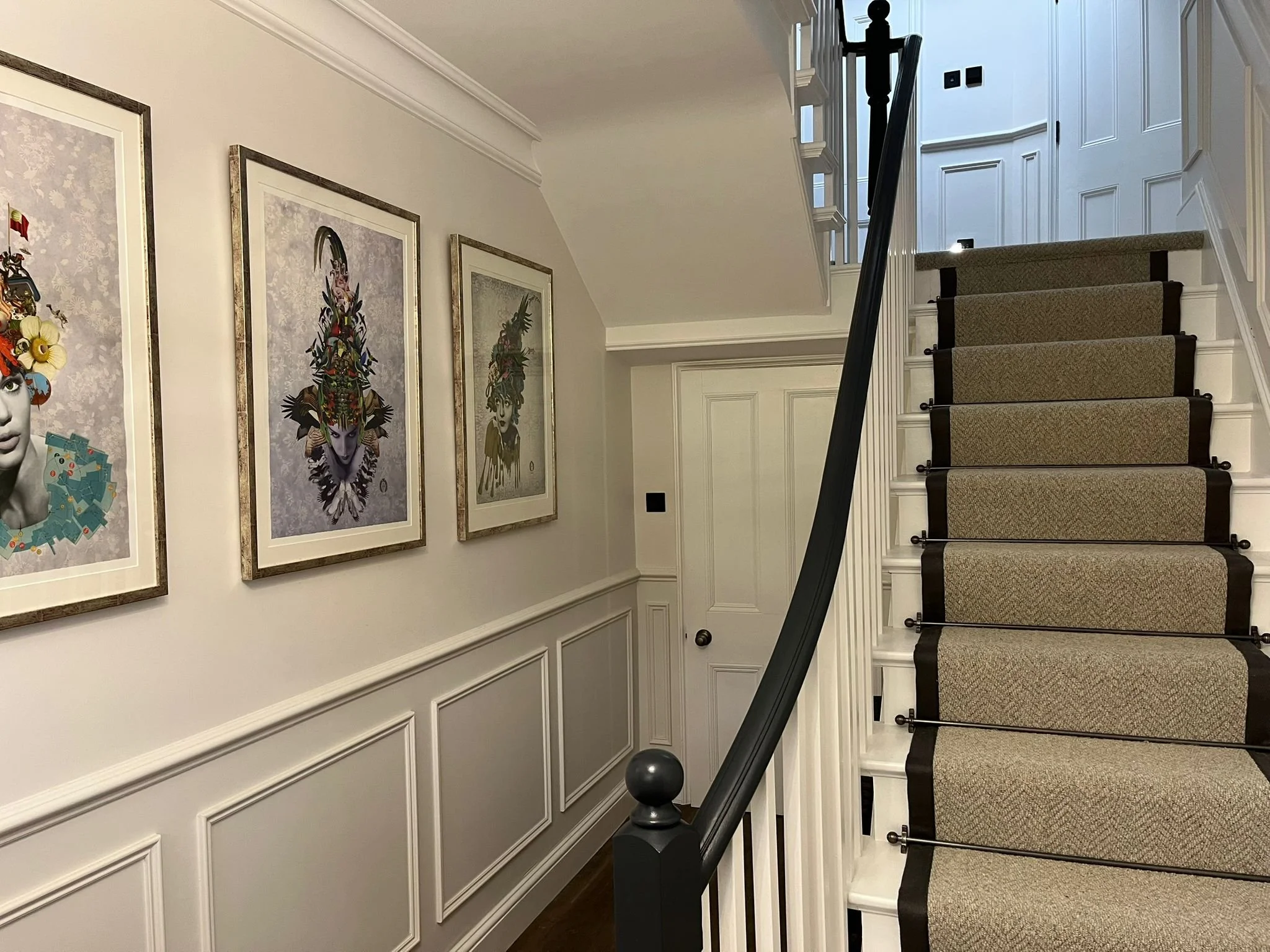 Interior of a house with a staircase, four framed artworks of stylized faces hanging on the wall, and a white wainscoting. The stairs are carpeted with a patterned runner and black stair rods.