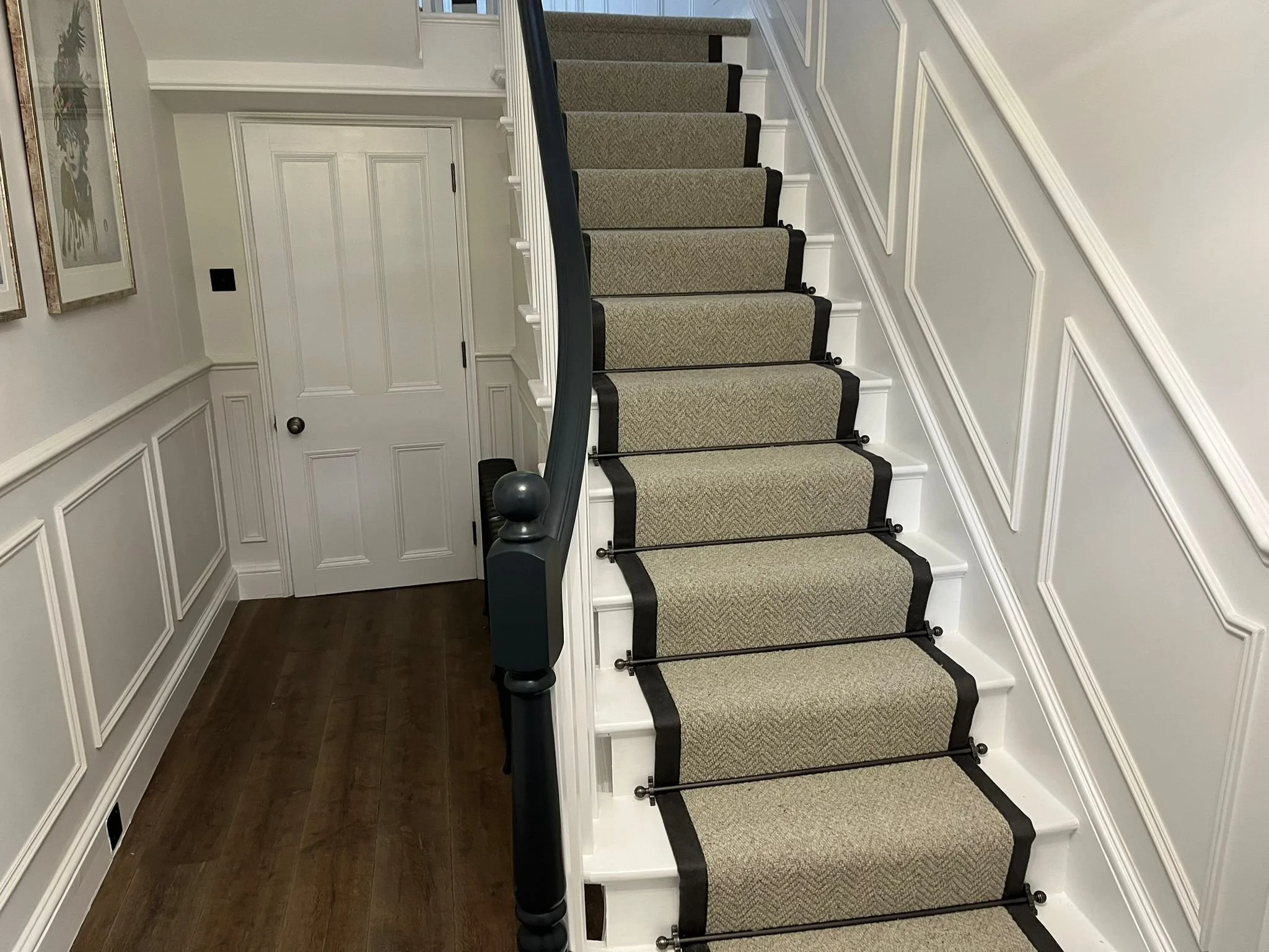 Interior view of a staircase with beige carpeted steps bordered in black, black handrail on the left, and white paneled walls. To the left, there is a corner with a dark wooden floor, a white door, and framed artwork on the wall.