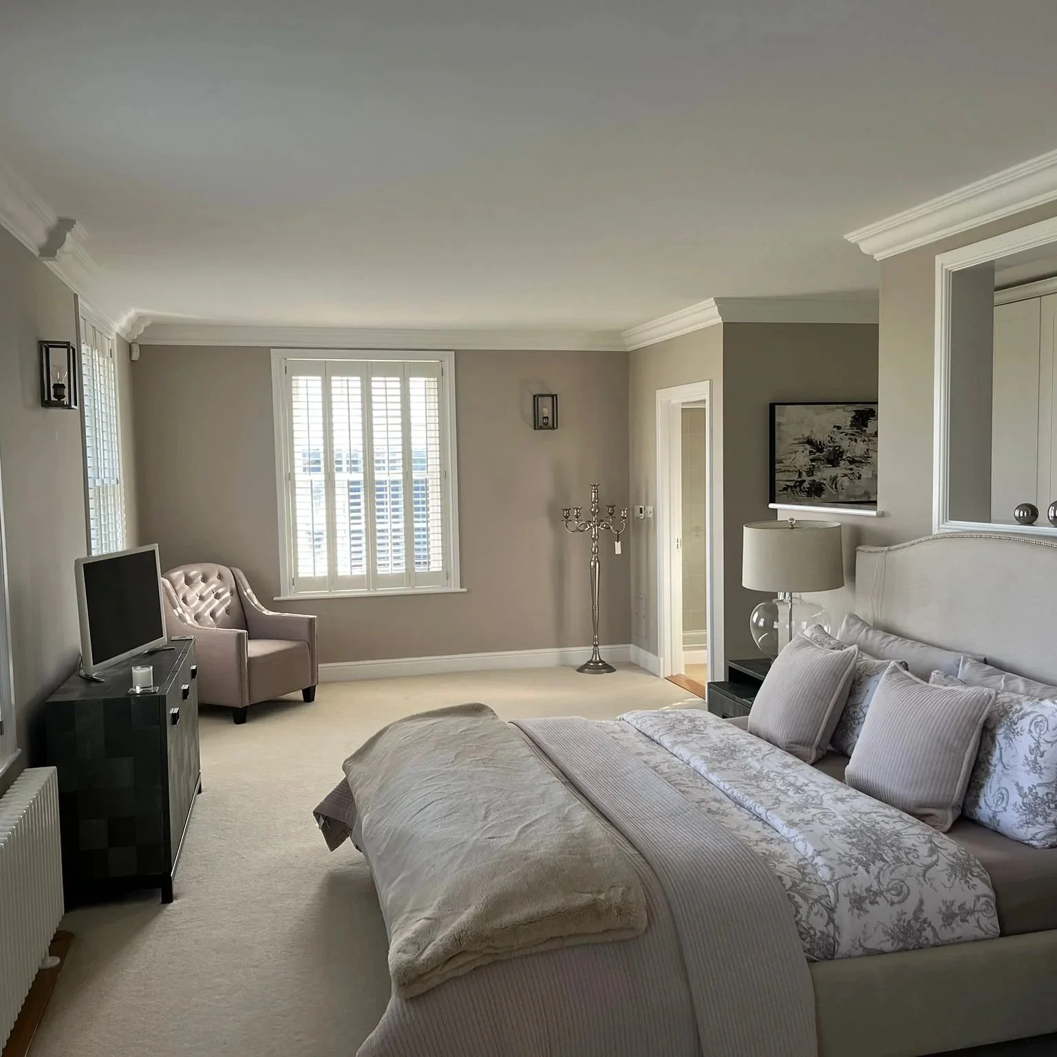 A spacious bedroom with a large bed, decorative pillows, and a headboard. There is a nightstand with a lamp, a TV on a stand, a purple armchair near a window with blinds, and wall-mounted lights. The room has beige walls and white crown molding.