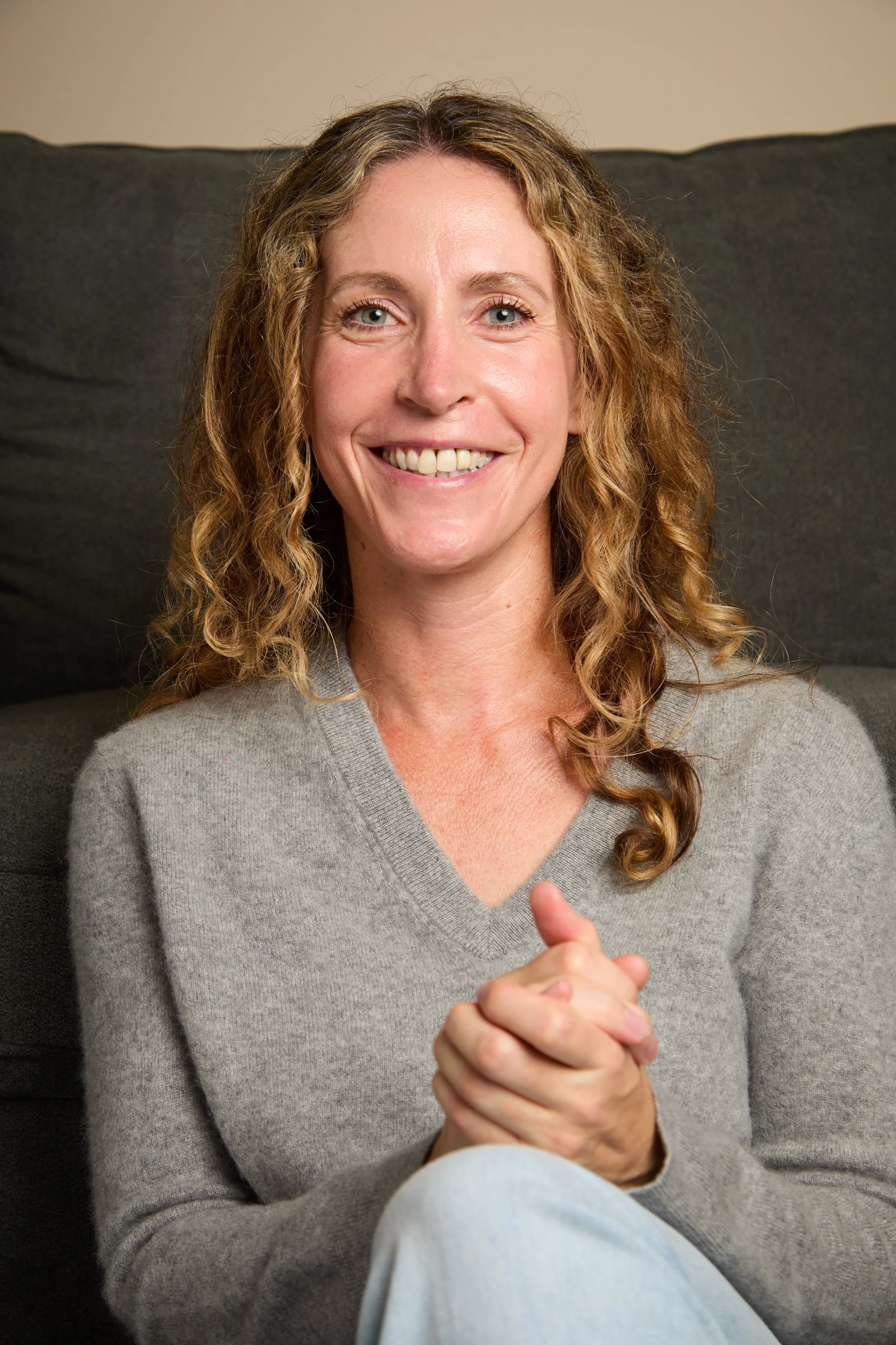 A smiling woman with curly blonde hair wearing a gray sweater, sitting on a black couch, with her hands clasped together in front of her.