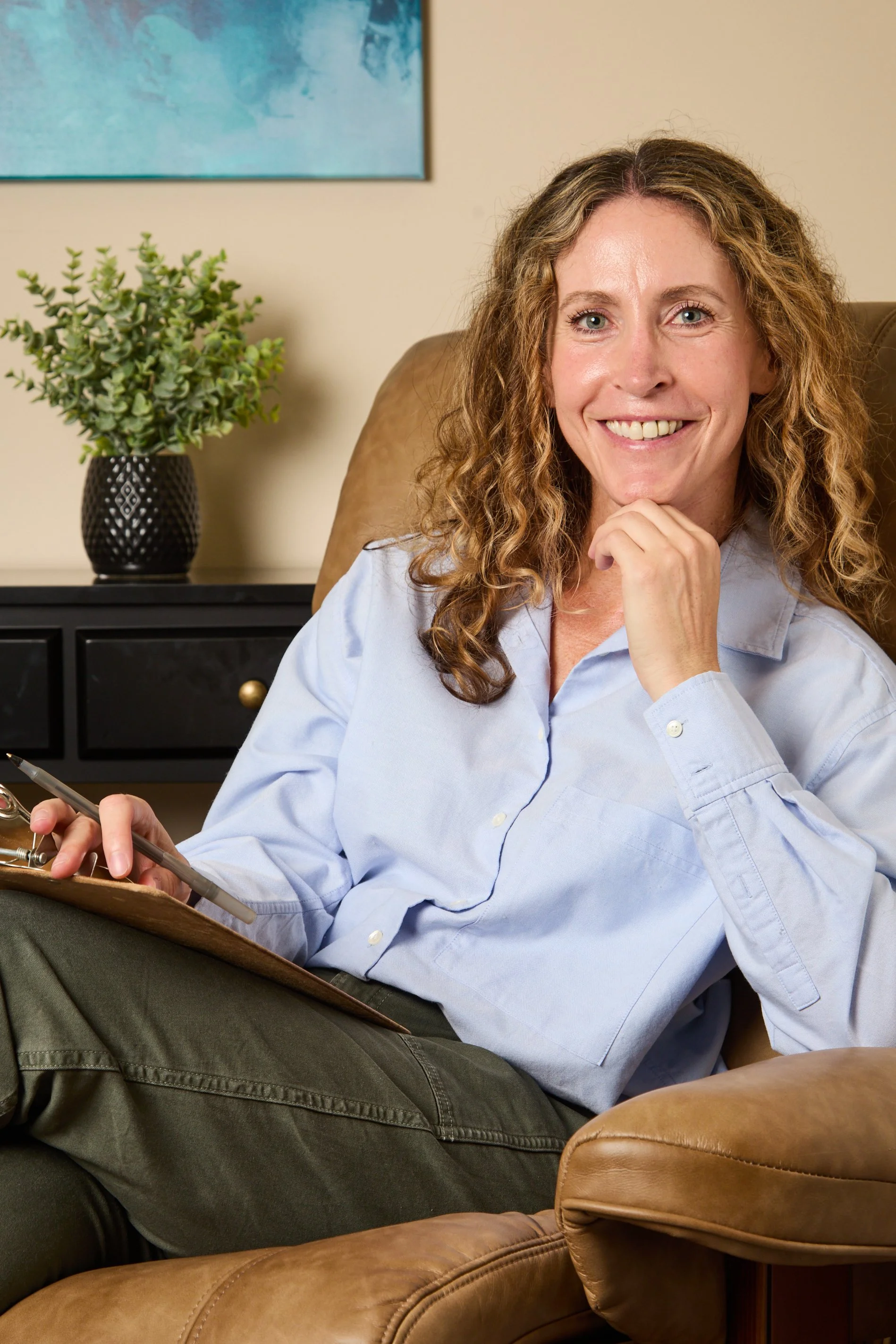 A woman with curly blonde hair and a light blue shirt sitting on a tan leather chair, smiling and holding a clipboard and pen, with a portrait on the wall and a potted plant on a black side table in the background.