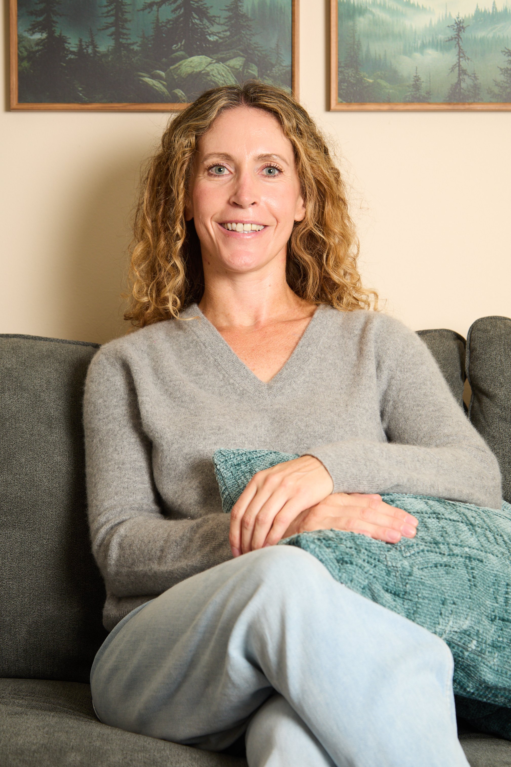A woman with curly blonde hair smiling, sitting on a grey sofa with a teal pillow, wearing a light grey sweater and light grey sweatpants, with framed nature landscape paintings of forest scenes hanging on the wall behind her.
