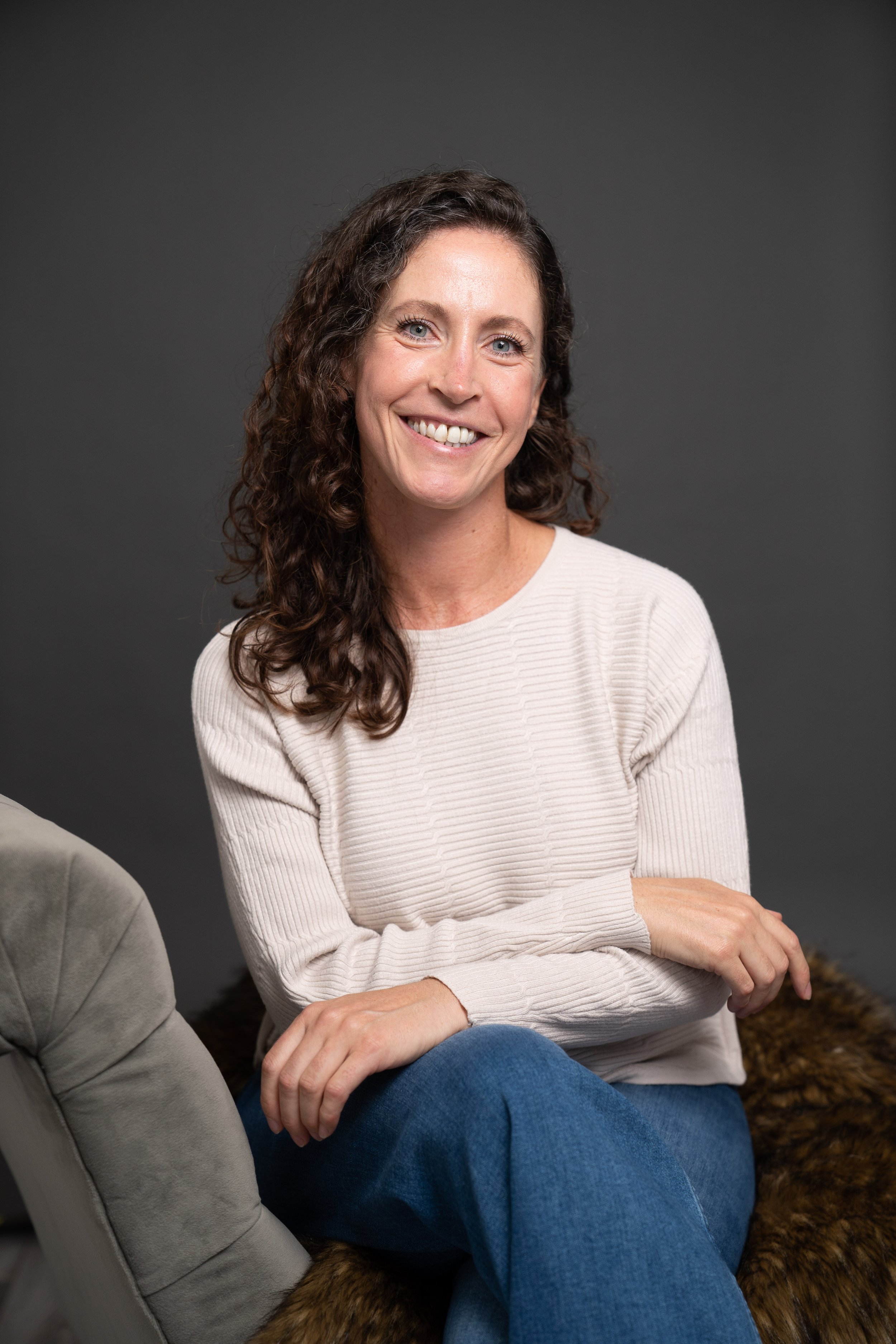 Portrait of a smiling woman with curly brown hair, wearing a white sweater and blue jeans, sitting on a fur-covered surface against a dark gray background.