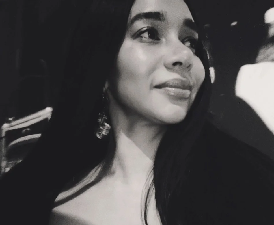 A woman with long dark hair and earrings is looking to the right, smiling gently in a black and white photo.