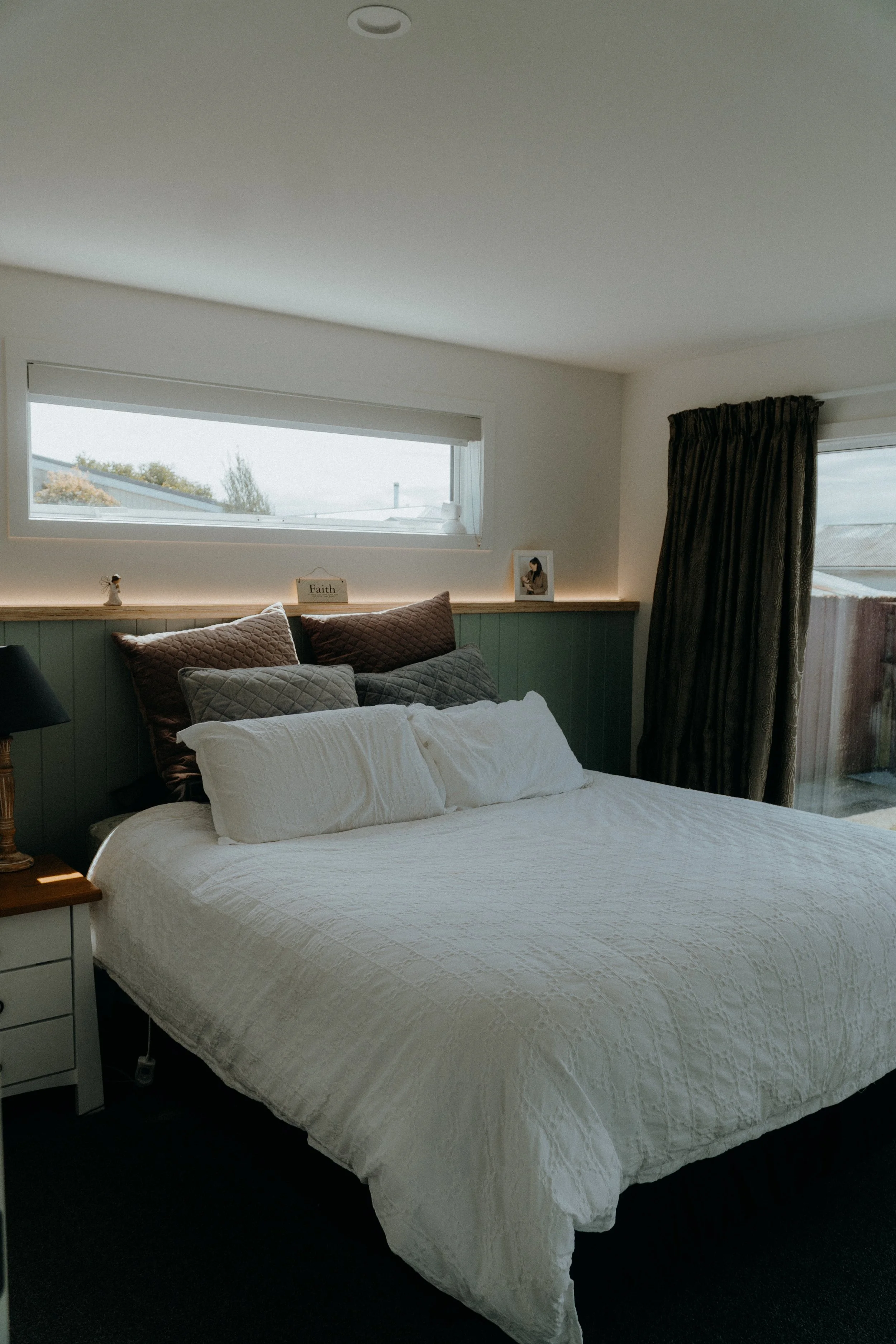 A neatly made bed with white bedding and multiple pillows in various shades of brown and gray in a cozy bedroom. There is a window behind the bed and a sliding glass door with curtains to the right.