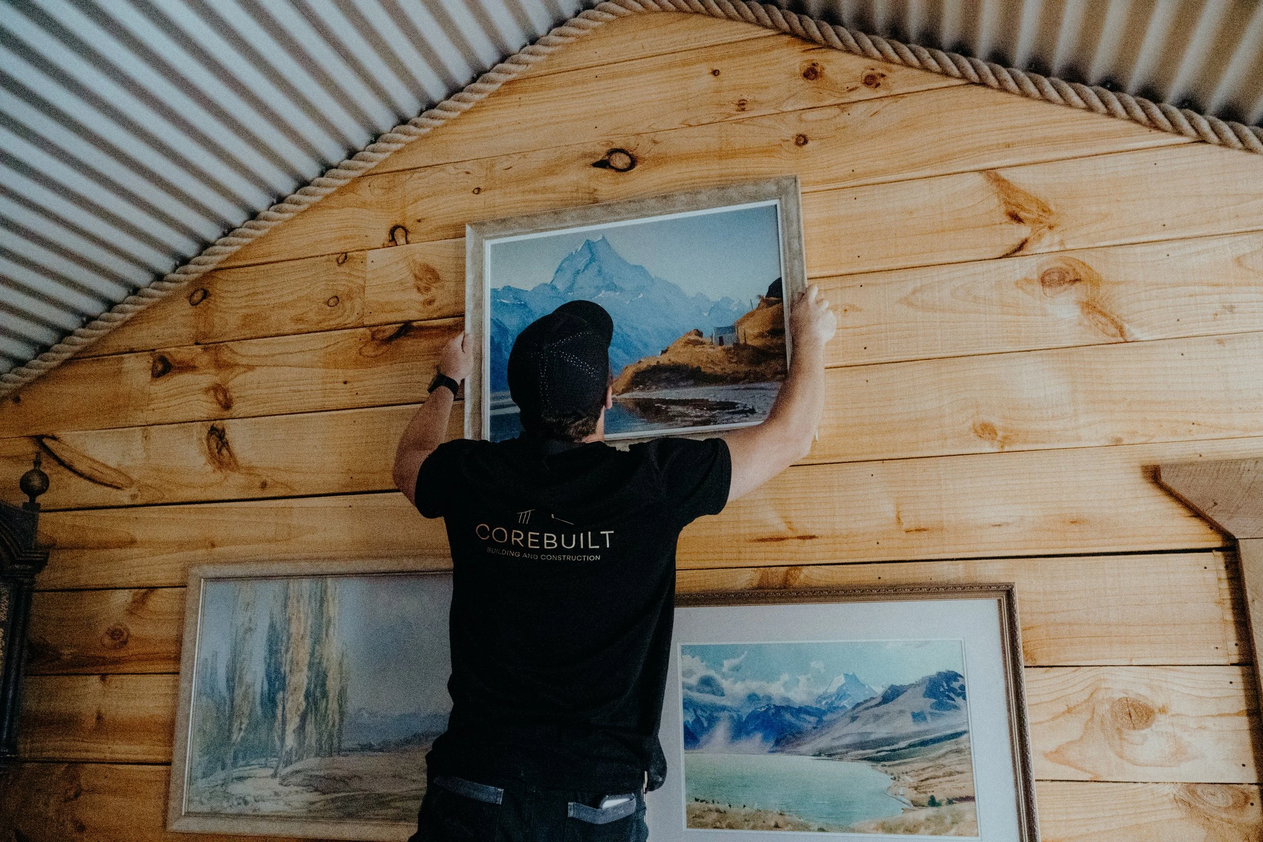 A person hanging a framed landscape painting of mountains on a wooden wall in an art gallery or studio.