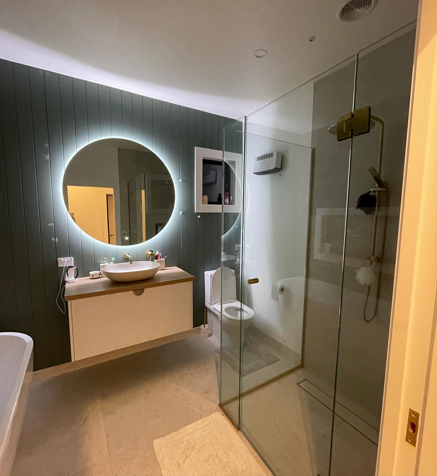 Modern bathroom with a backlit round mirror above a vessel sink on a wooden vanity, a white toilet, a glass shower enclosure, and a dark accent wall with vertical paneling.