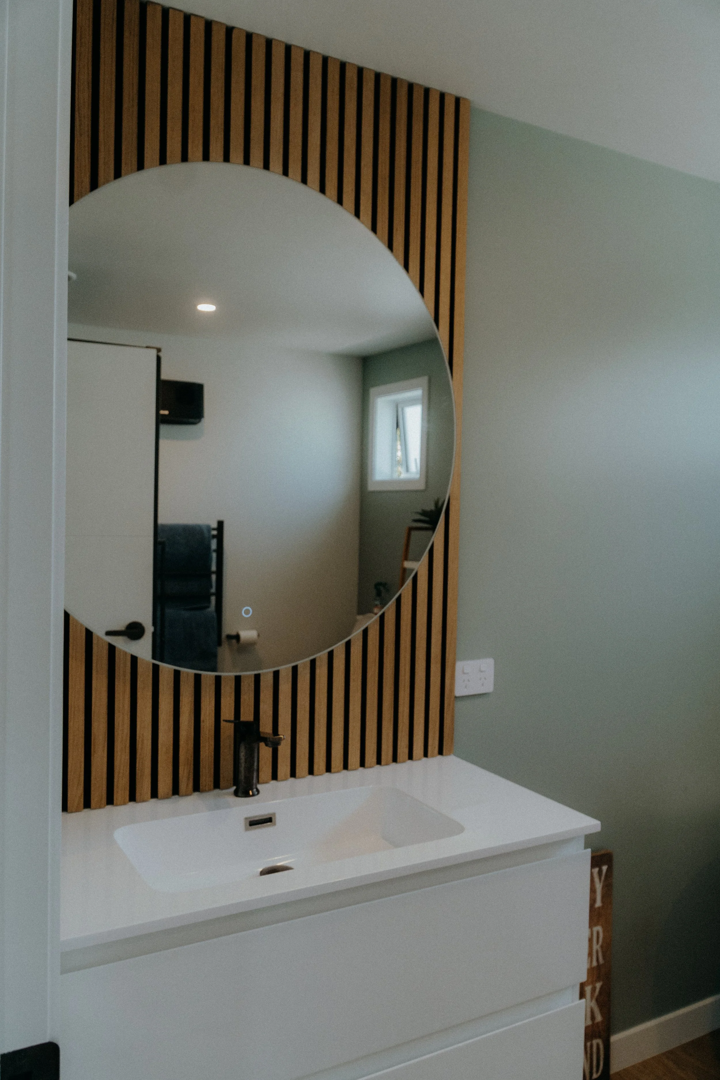 Modern bathroom with a white sink, black faucet, round mirror, and wooden slat wall panel behind the mirror.