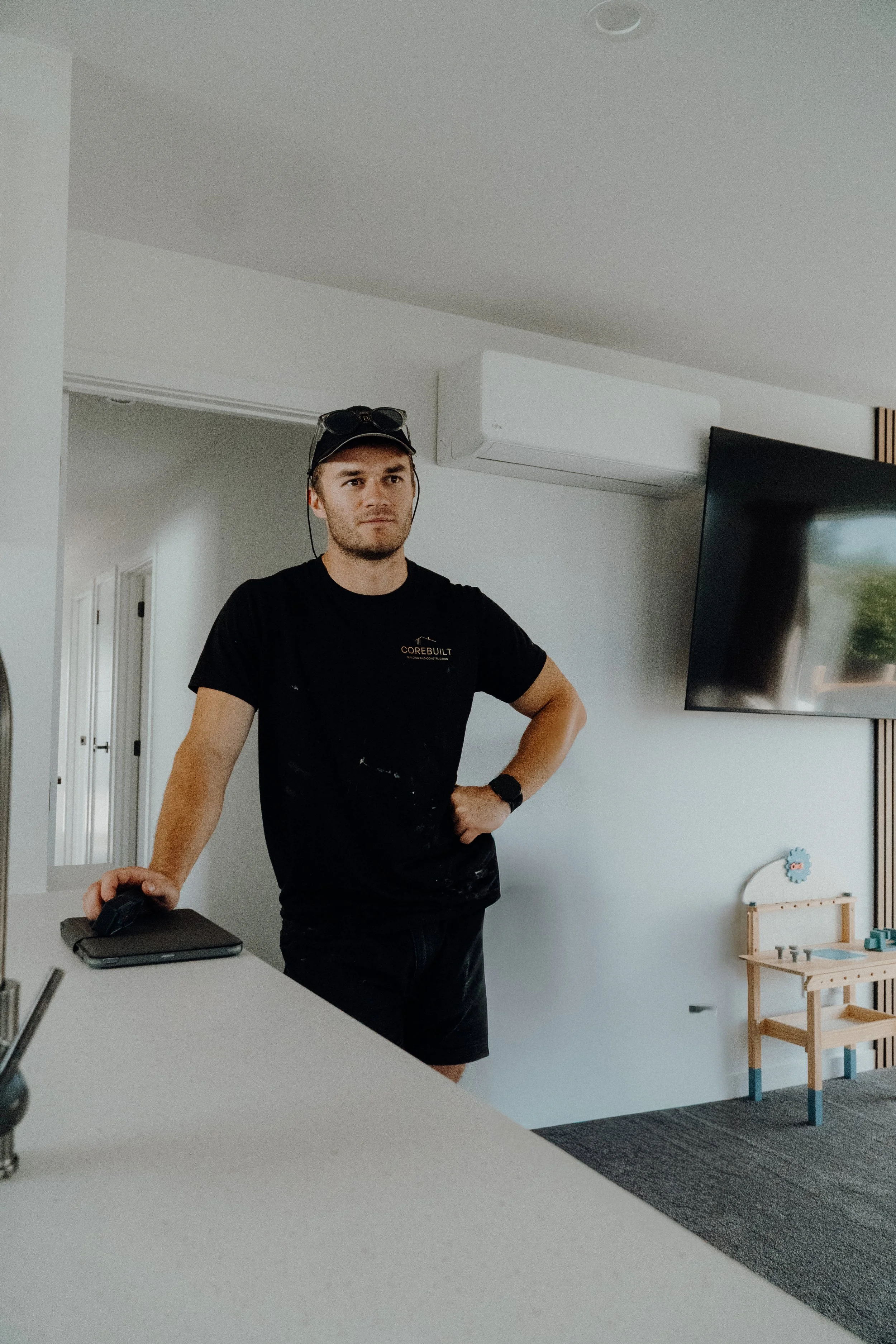A man wearing a black t-shirt and shorts, standing with one hand on his hip and the other on a countertop, in a modern room with a wall-mounted TV and a children’s play area.