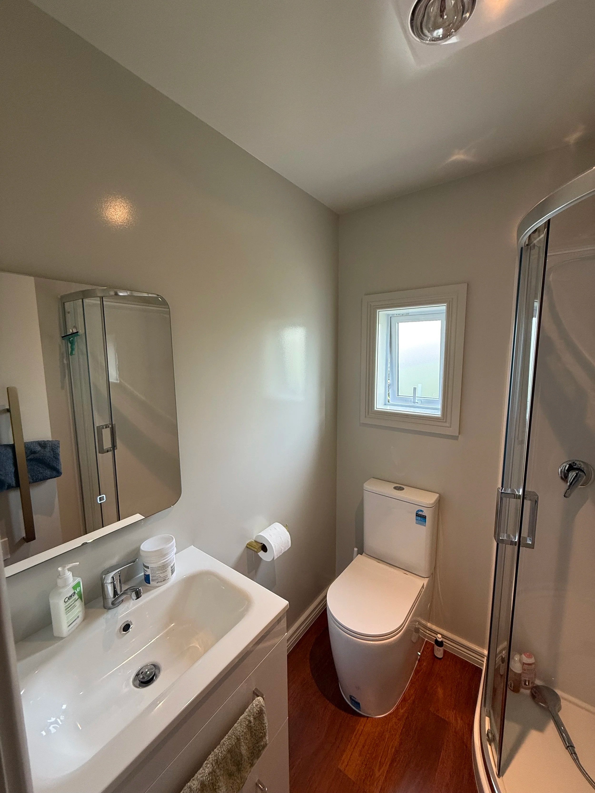 A small bathroom with a white sink and mirror, a toilet, a small window, and a corner shower with a glass door.