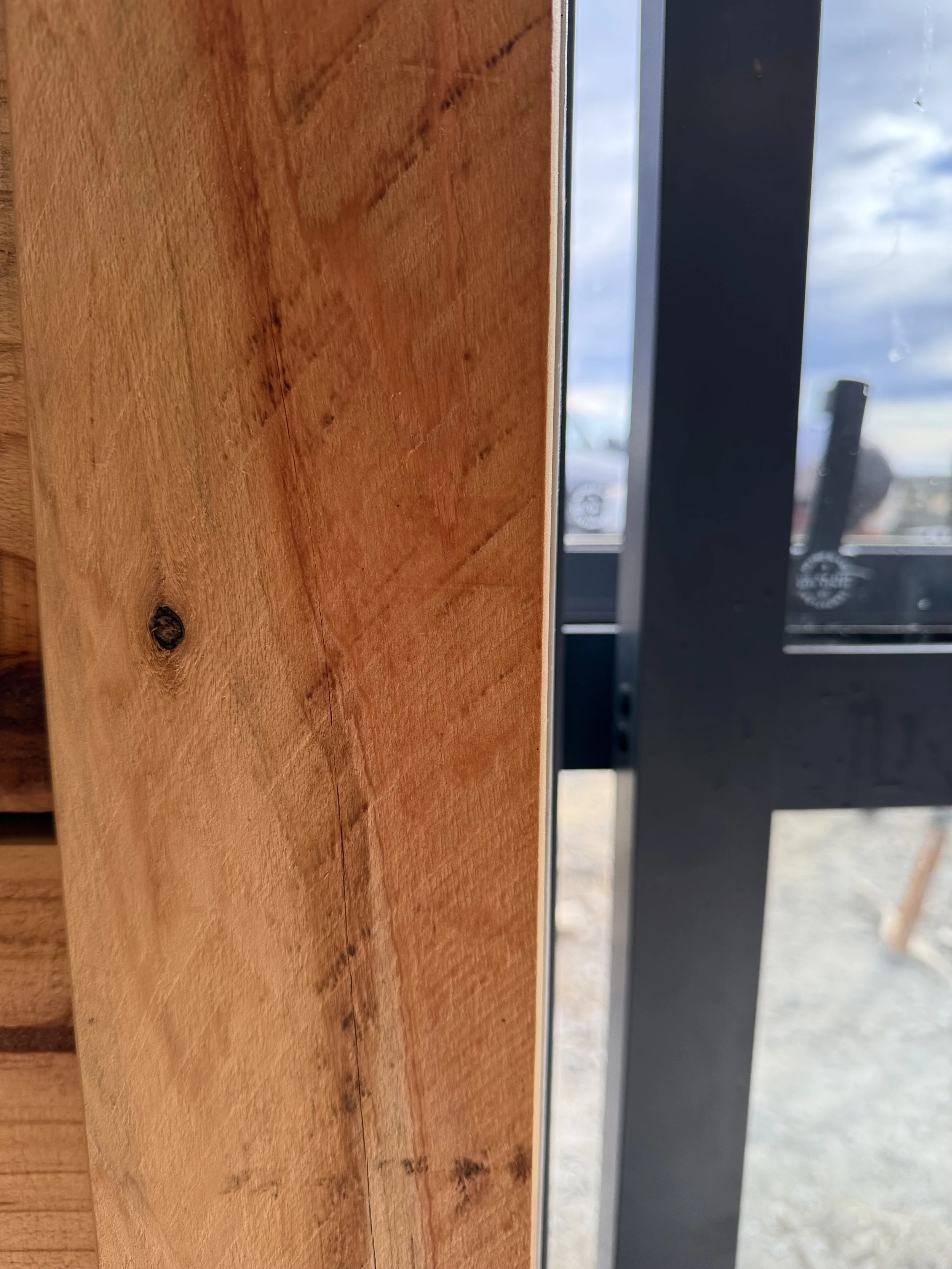 Close-up of a wooden wall with a corner next to a window showing outdoor sky and clouds.