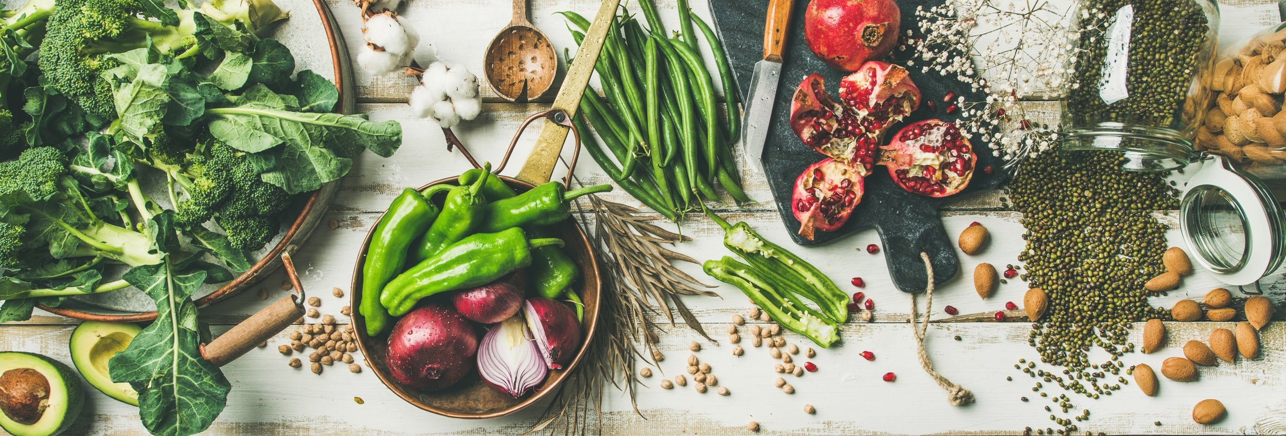 Fresh vegetables and fruits on a rustic wooden table, including broccoli, avocados, onions, green peppers, pomegranate, green beans, and various jars of legumes and spices.