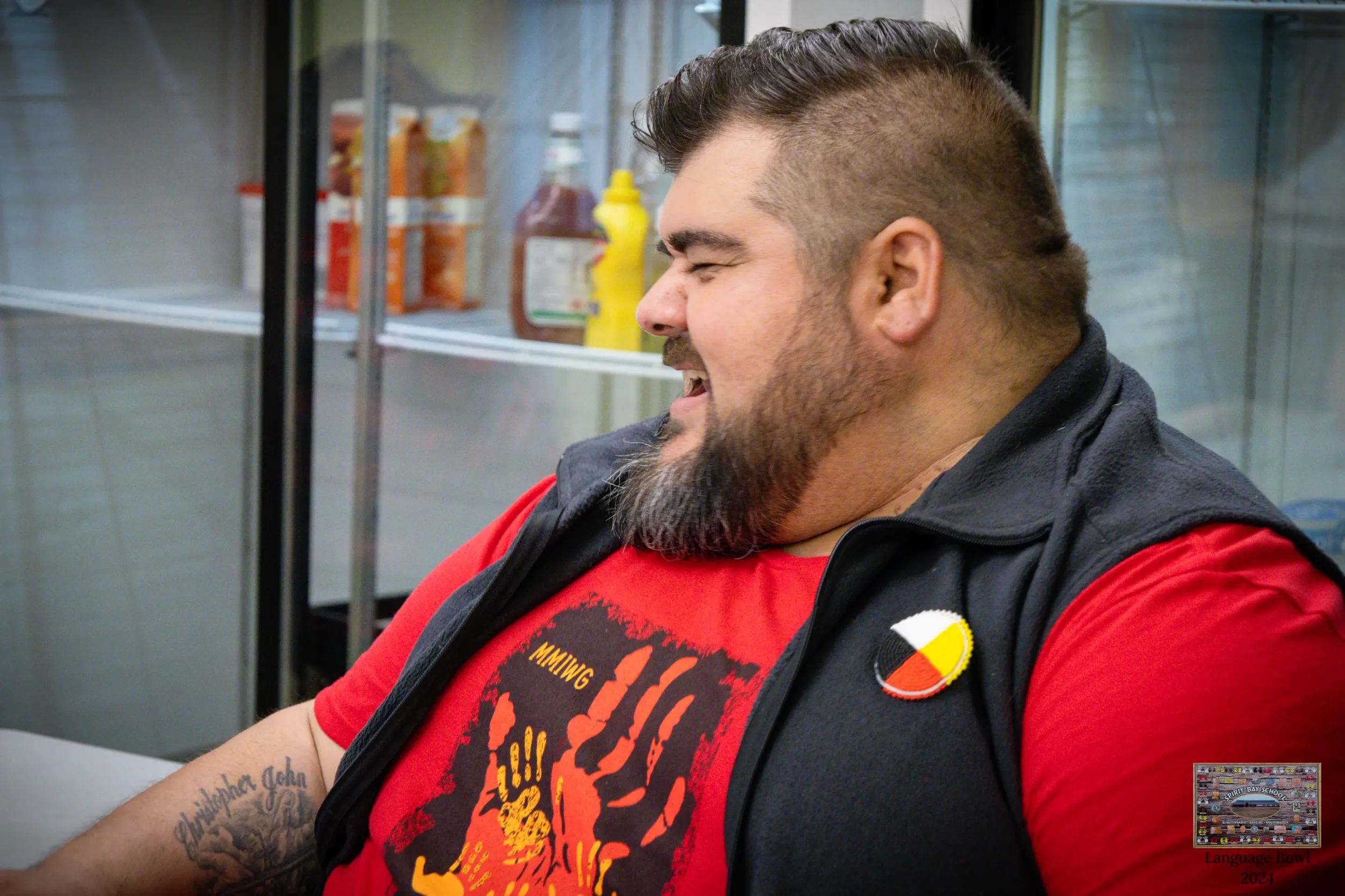 A man with a beard and mustache, wearing a red t-shirt and a black vest with a badge, sitting in front of a refrigerator in a casual setting, smiling with eyes closed.