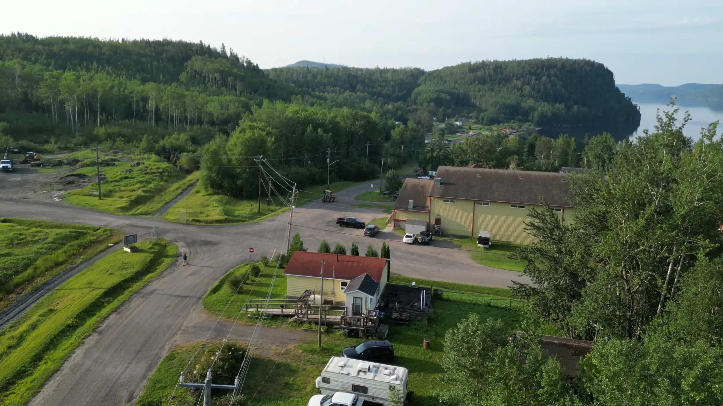 Aerial view of a small town near a large body of water with forested hills in the background. There are several houses, parked cars, power lines, and a gravel road.