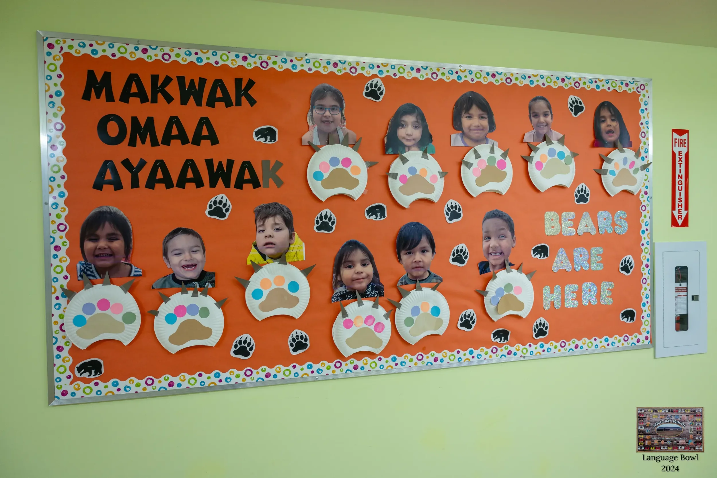 Classroom bulletin board with children's photos and bear paw prints, featuring the phrase 'Makwak Omaa Ayaa Wk Bears Are Here'. The board is decorated with colorful borders and is on a light green wall.