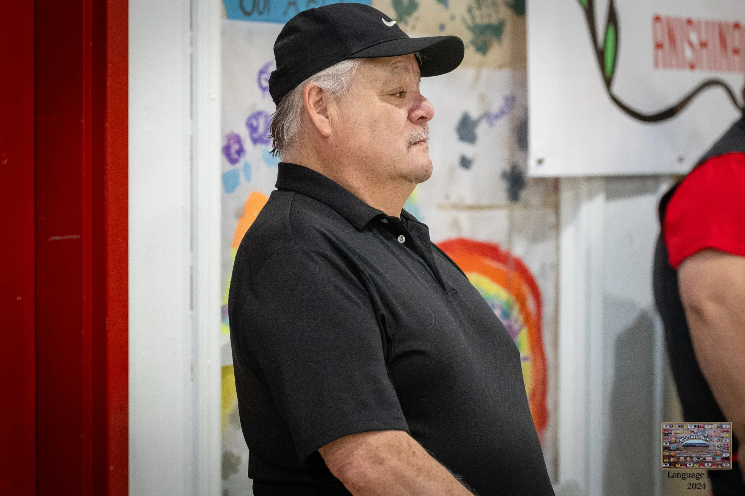 A man with gray hair wearing a black Nike baseball cap and black polo shirt standing indoors with colorful children's artwork in the background.