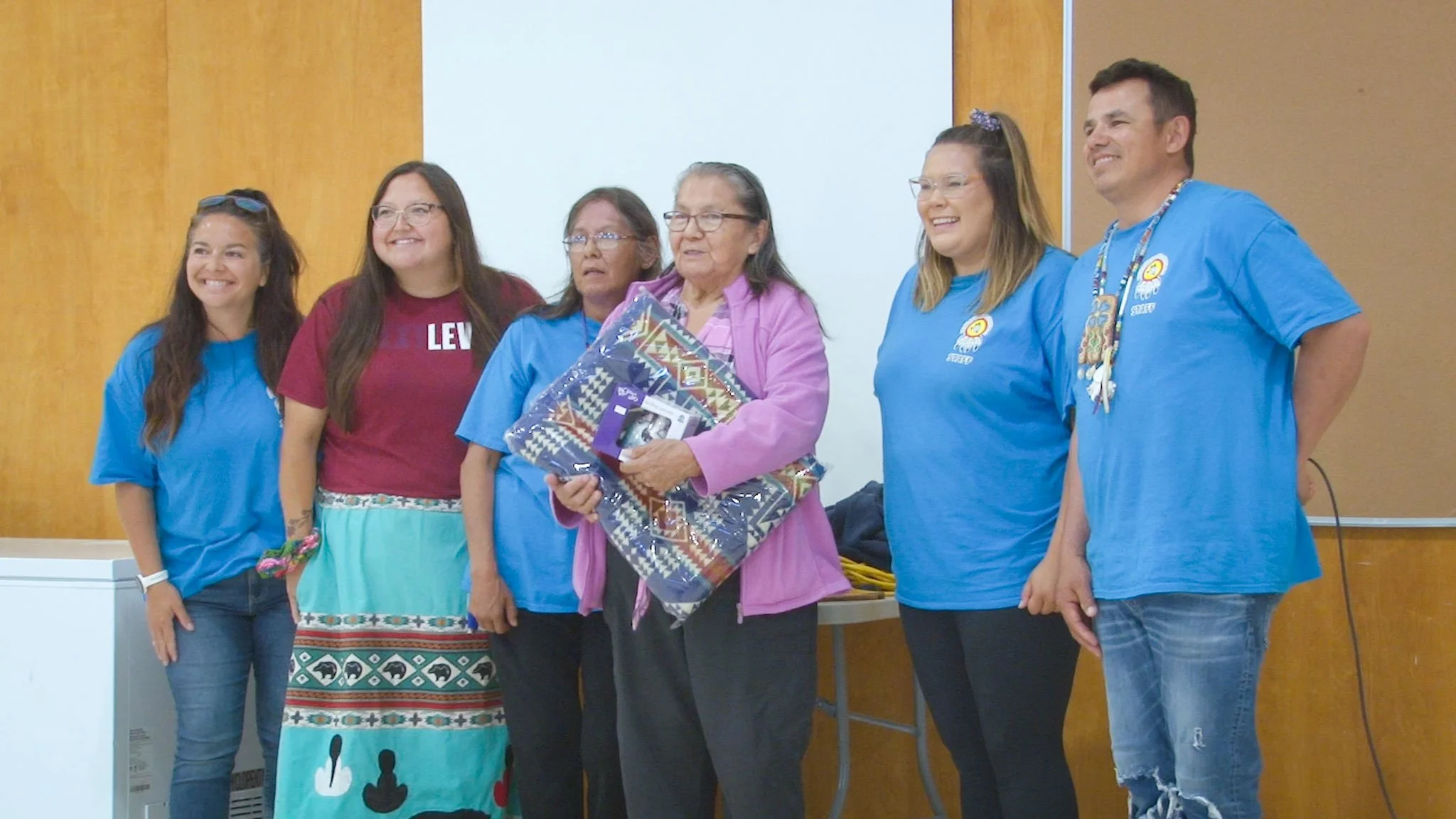Six people standing in a room, with three women wearing blue shirts, one woman in a maroon shirt, and an elderly woman holding a gift, smiling for a photo.