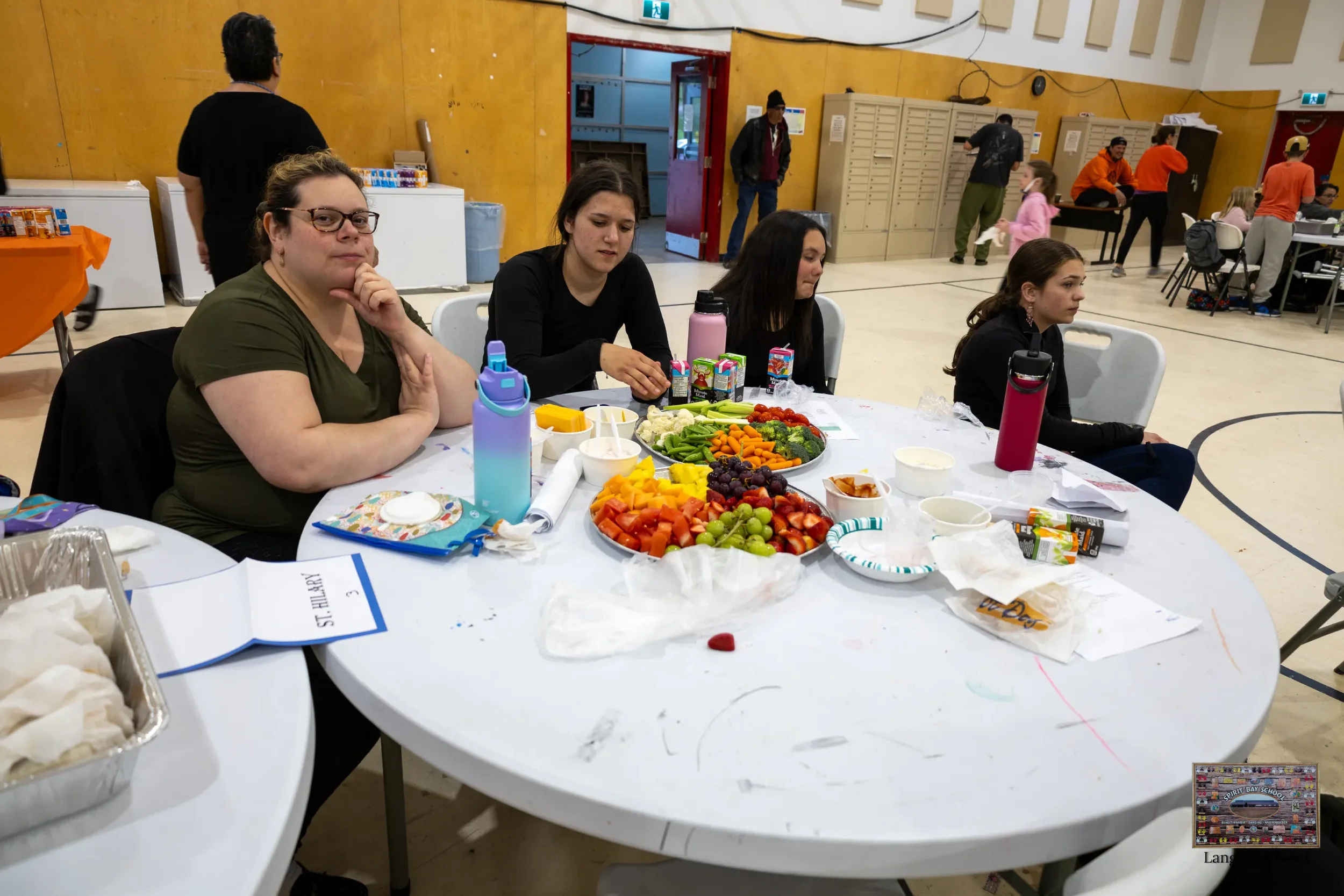 People sitting around a table with a variety of colorful fruits and snacks in a gymnasium or community center.