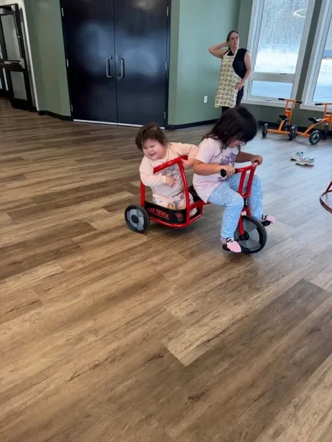 Two young girls are riding in a red and black toy pedal truck inside a room with wooden flooring. One girl is sitting in the back, smiling, and the other girl is pedaling in front. In the background, a woman is standing near a window, watching the children.
