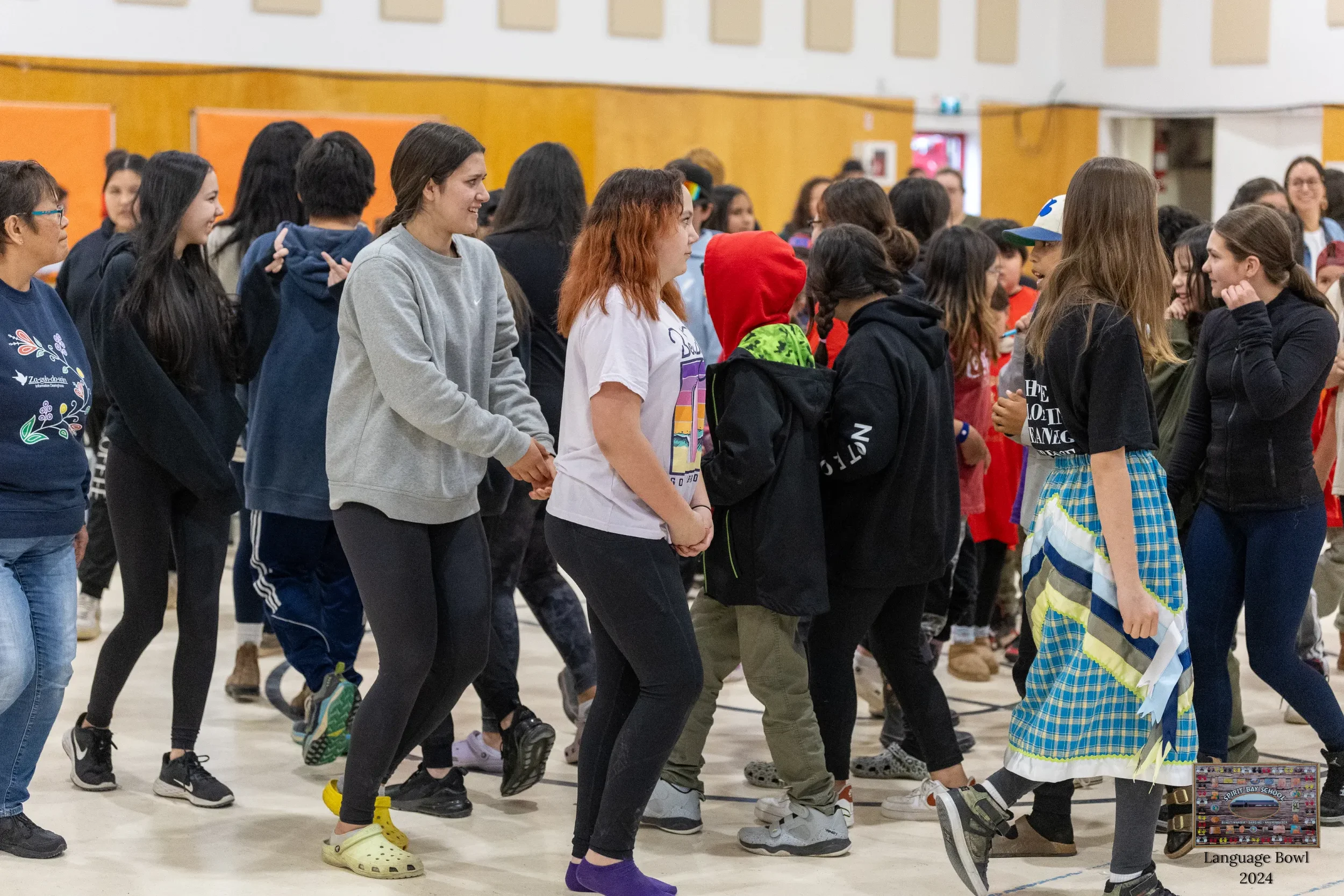 A group of students in a gymnasium participating in a dance or activity, some are holding hands, others are standing and watching, with diverse clothing including hoodies, t-shirts, and a traditional dress.