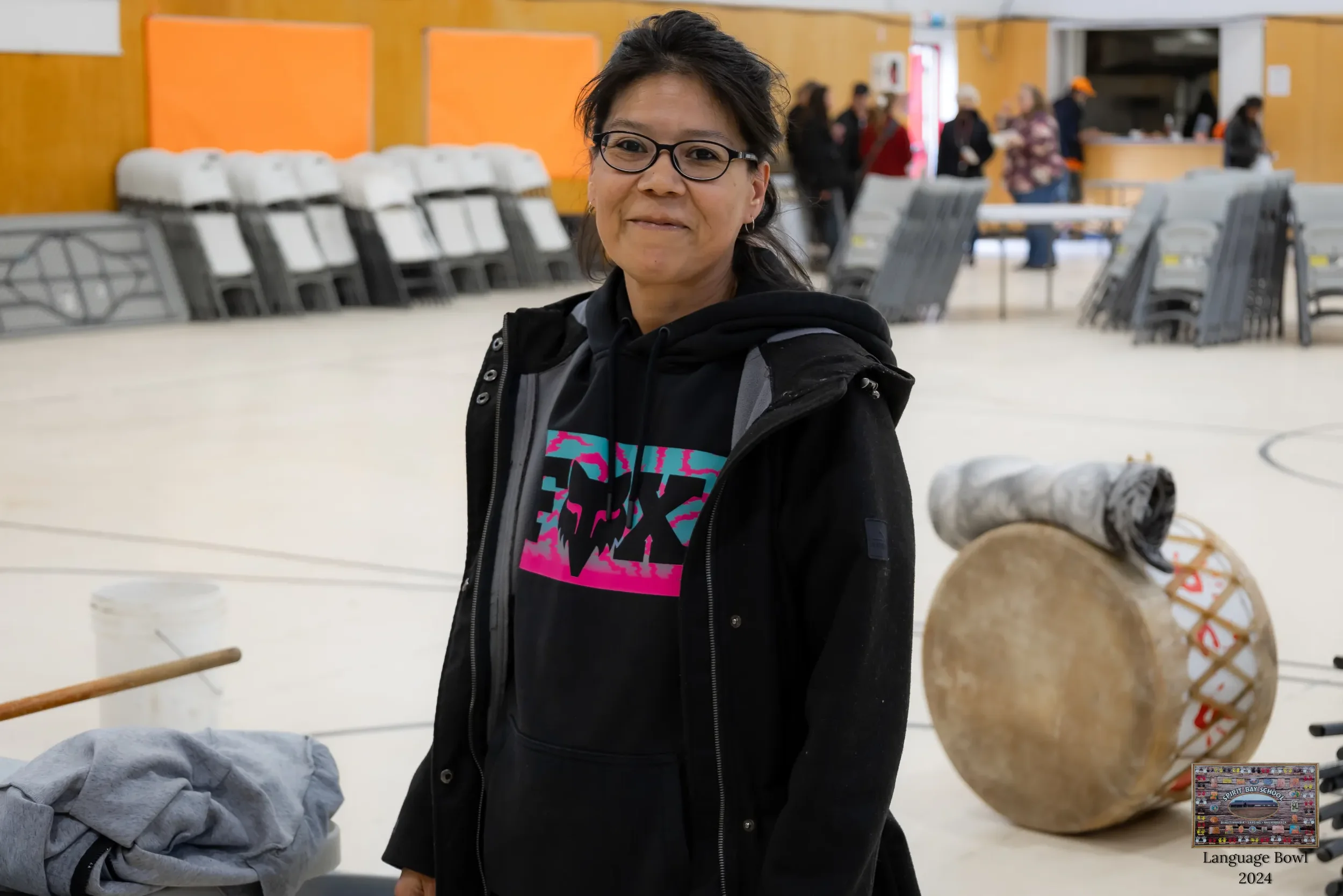 A woman with dark hair and glasses standing in an indoor gymnasium. She is wearing a black hoodie with a colorful graphic at the front and a black jacket. In the background, there are people, stacked chairs, and a large drum on the floor.