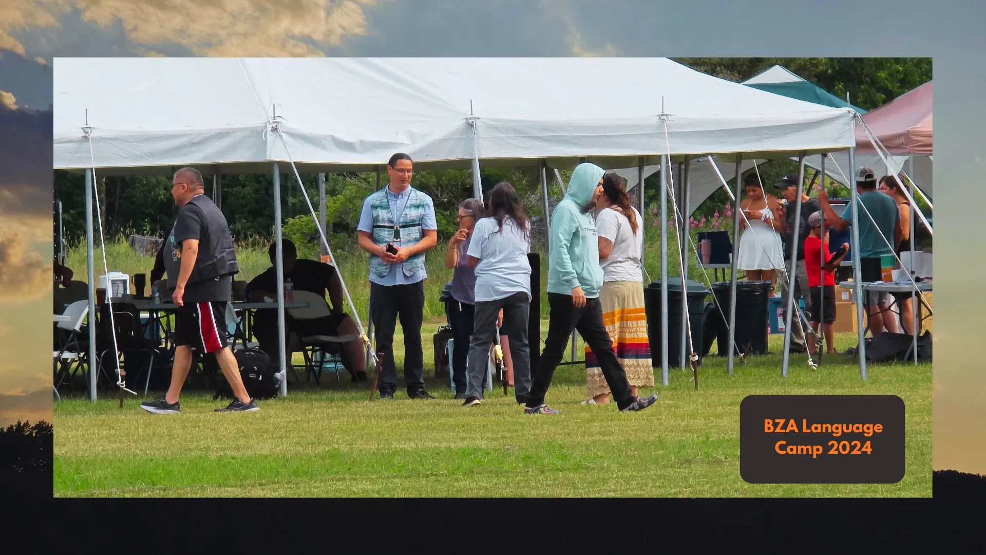A group of people gathered under a white tent outdoors on grass during daytime, at BZA Language Camp 2024, engaging in conversation and activities.