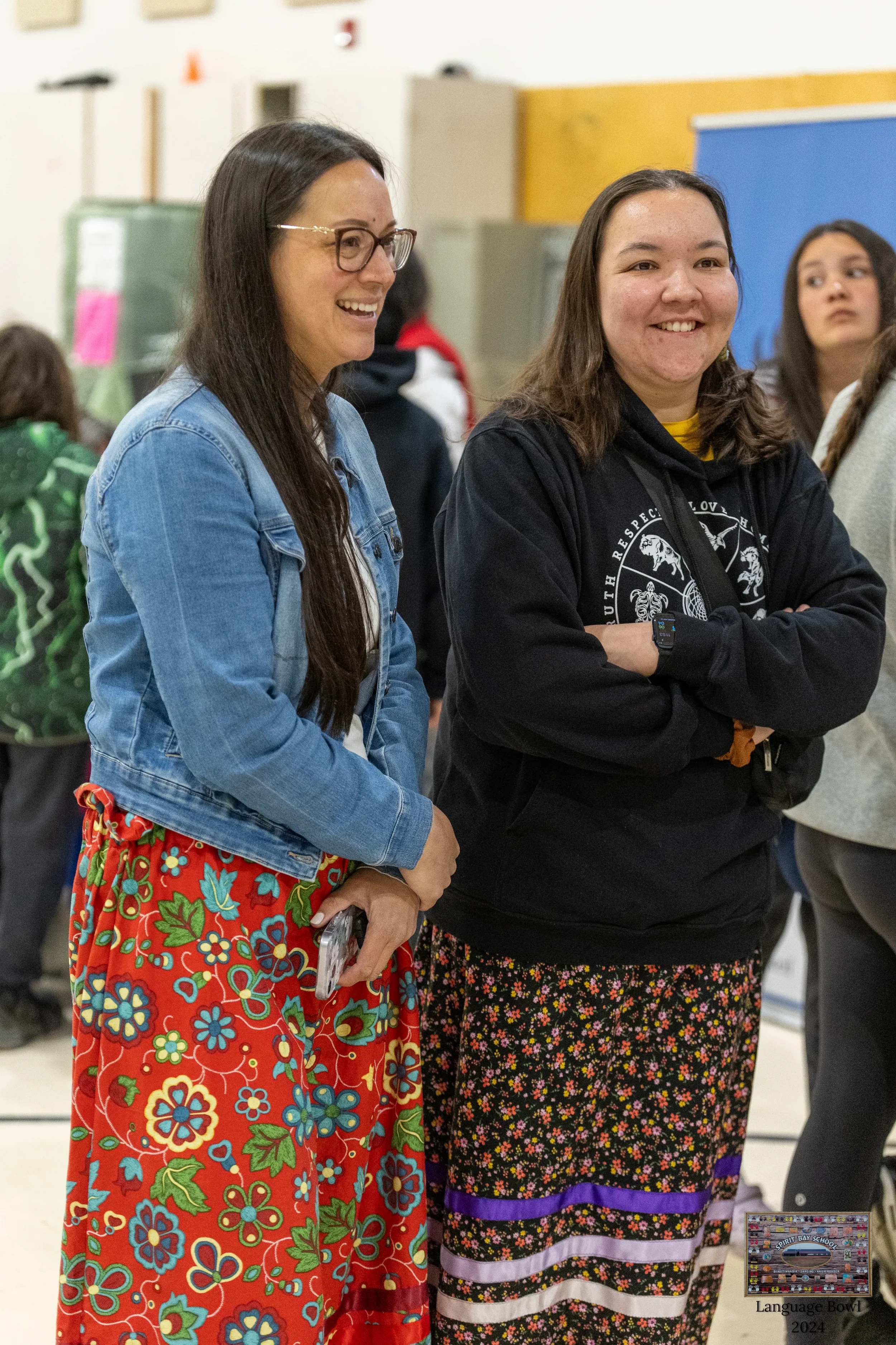 Two women standing and smiling at an indoor event, with other people in the background. One woman wears glasses, a denim jacket, and a red floral skirt. The other woman wears a black hoodie with a logo and a floral skirt with a purple ribbon.