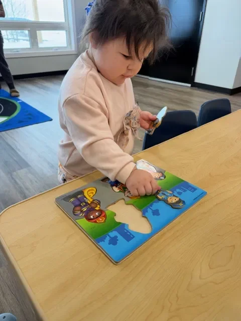 A young child with dark, tousled hair playing with a puzzle at a wooden table in a room with large windows and hardwood floors.