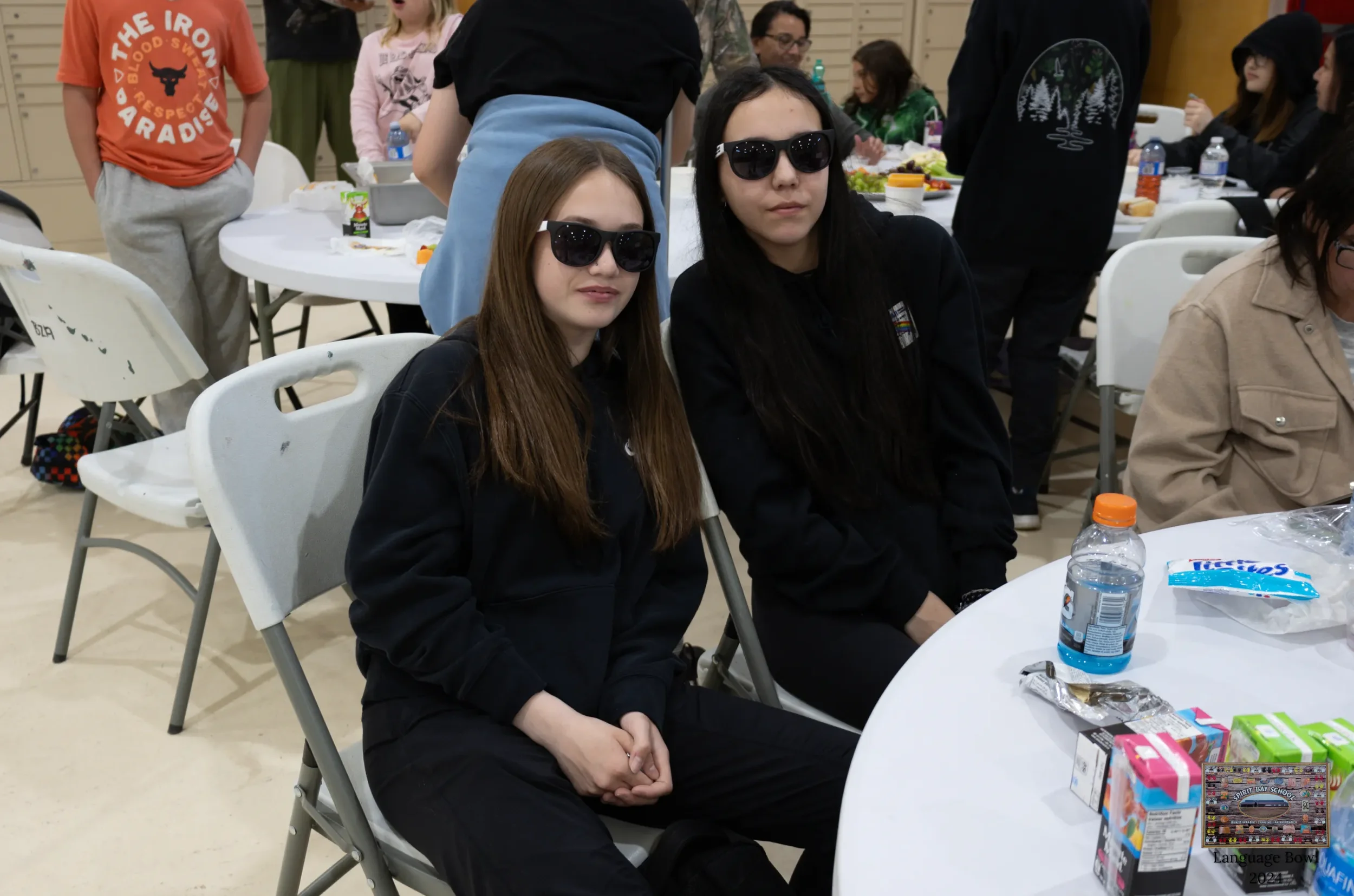 Two young women sitting at a round table wearing black hoodies and sunglasses, with multiple people in the background at an indoor gathering or party.