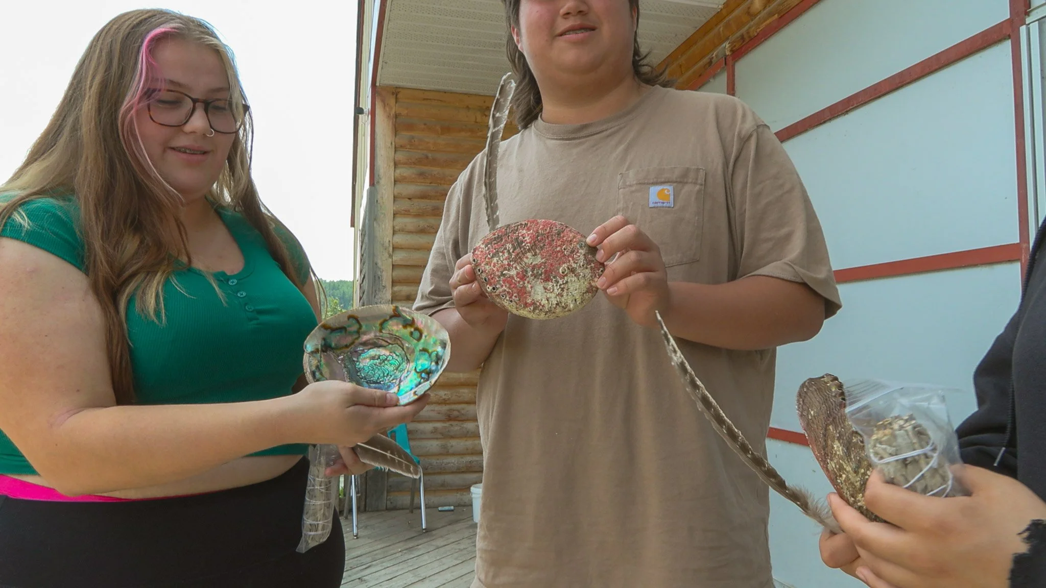 Three people holding colorful shells and a piece of driftwood outdoors near a wooden and metal building.