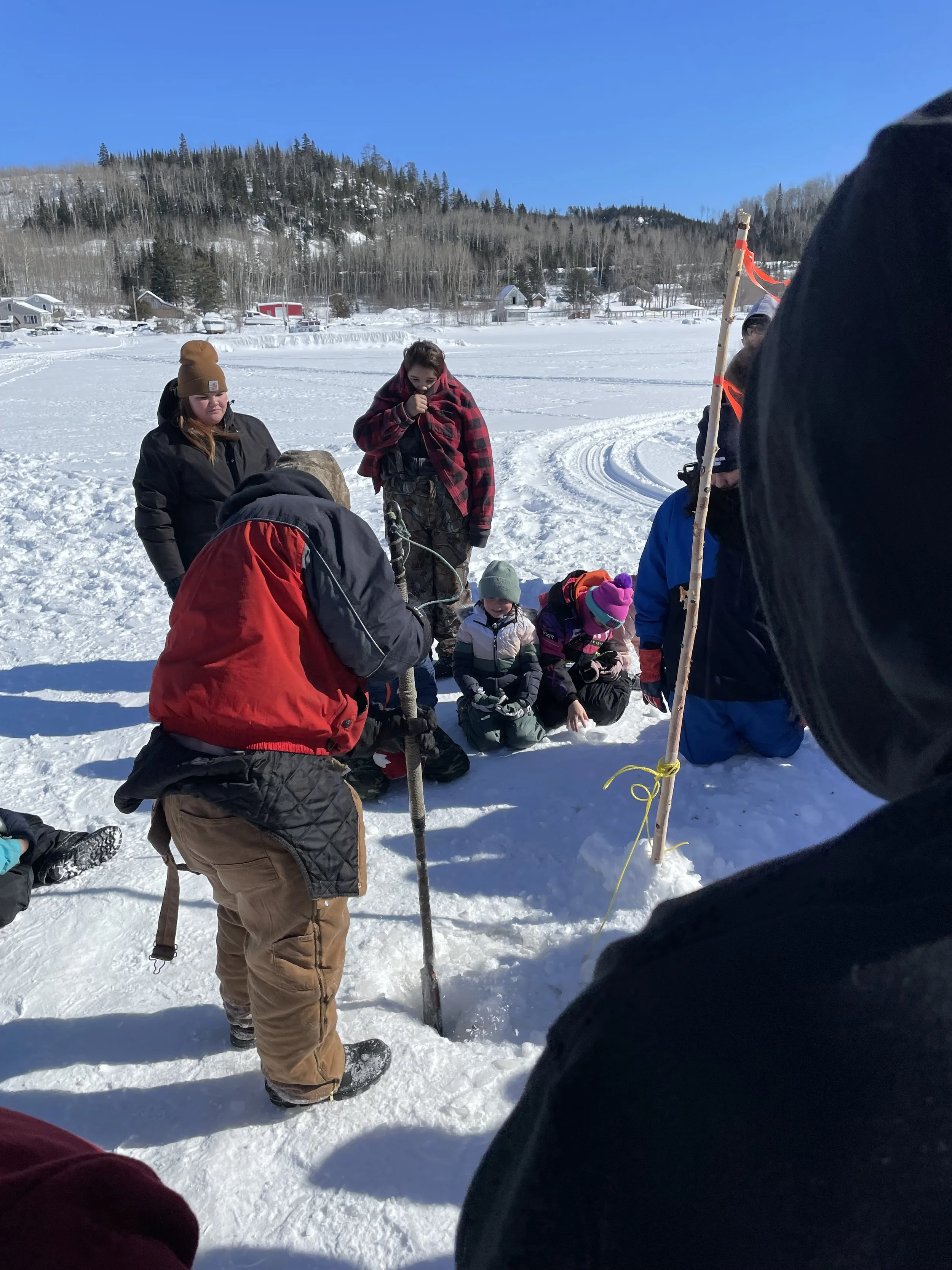 Group of people gathered on a snowy frozen lake, participating in an outdoor activity or lesson, with snow-covered trees and houses in the background under a clear blue sky.