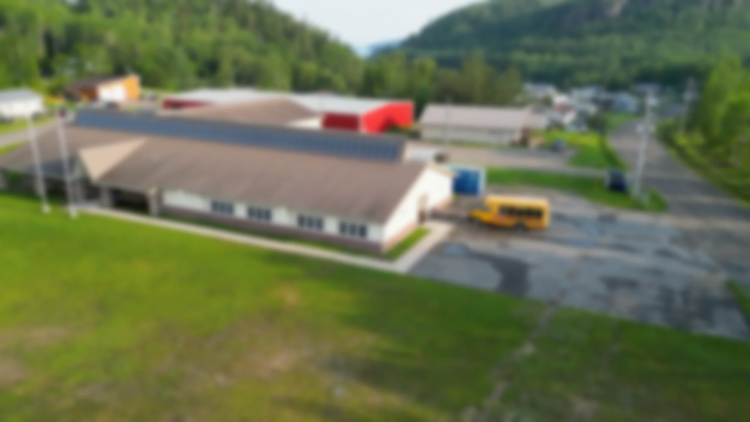 A blurry aerial view of a small building with a parking lot, school bus, and surrounding green landscape and trees.
