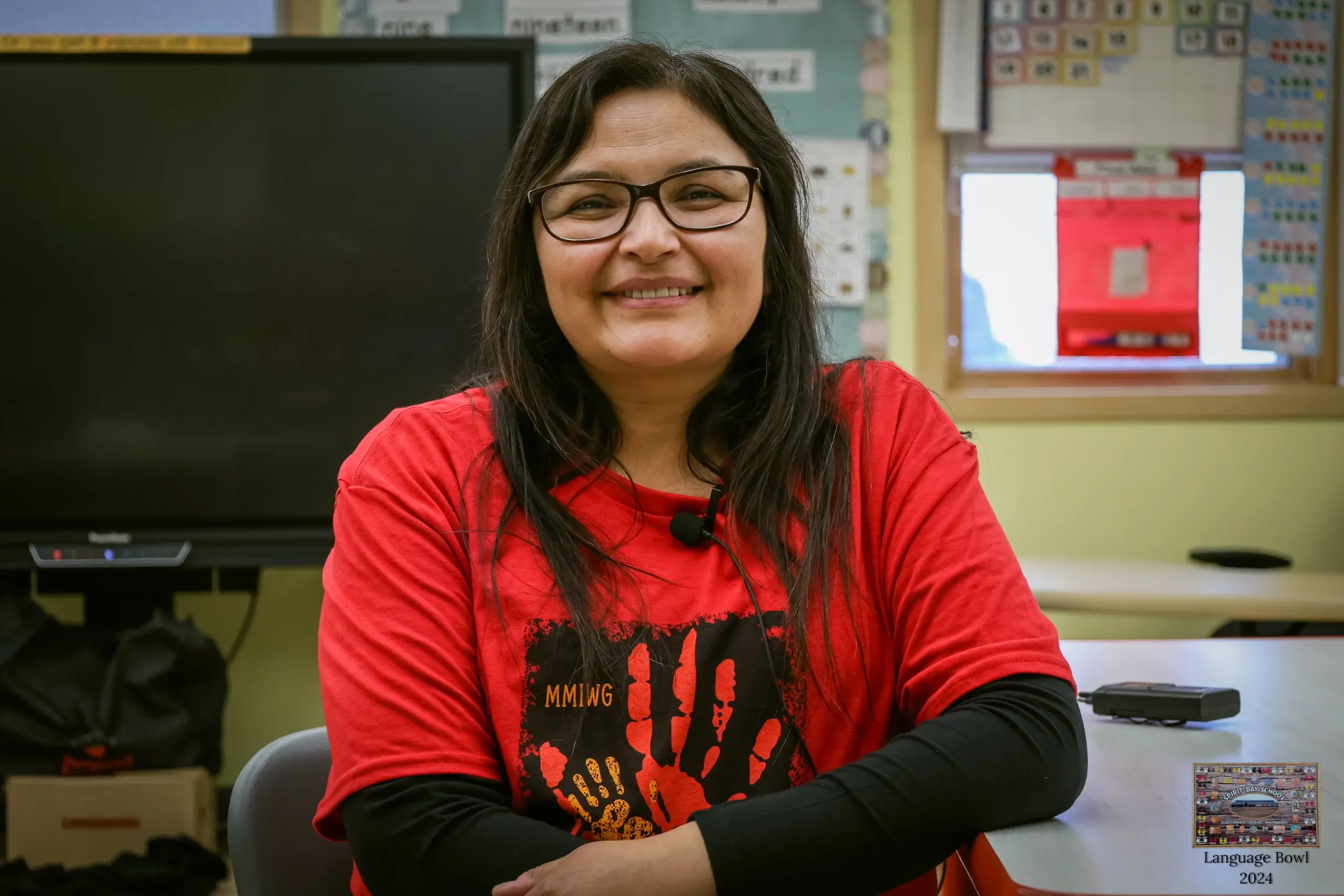 A woman with glasses and long dark hair, wearing a red t-shirt with black sleeves, smiling at the camera in a classroom setting.