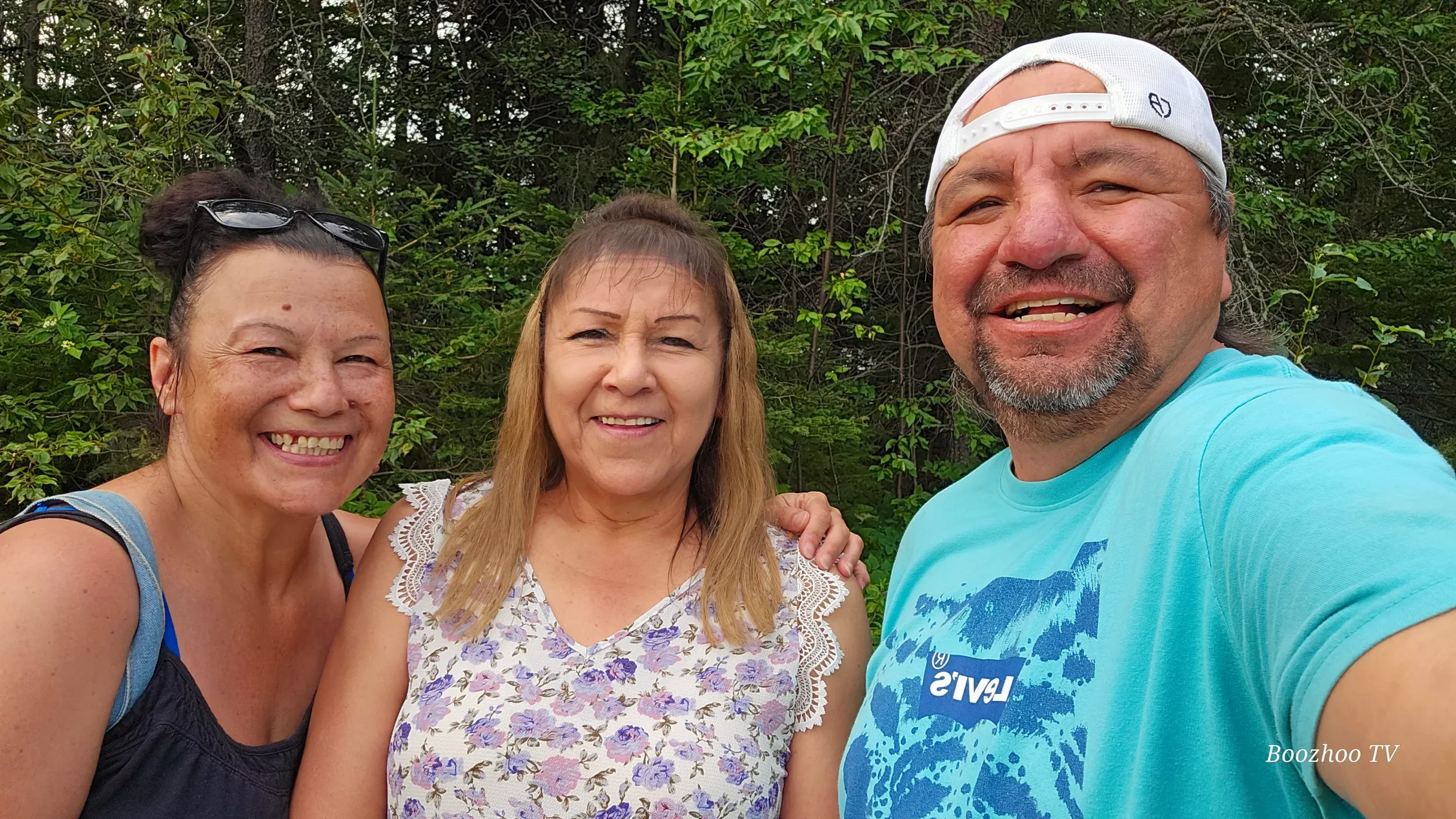 Three smiling people taking a selfie outdoors in front of green trees, two women and one man, all casually dressed.