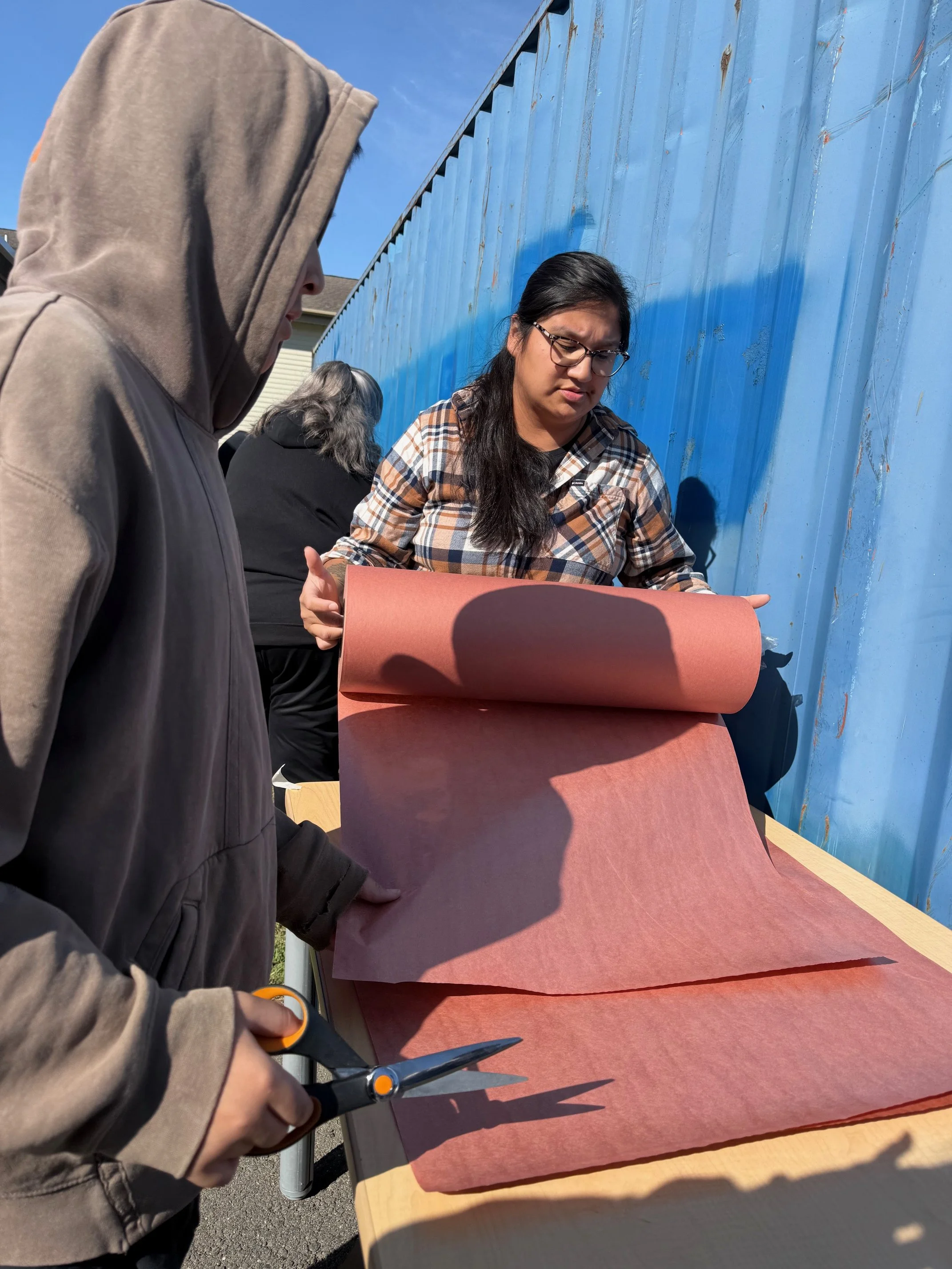 People working with large sheets of colored paper outdoors on a table, with a blue fence in the background.