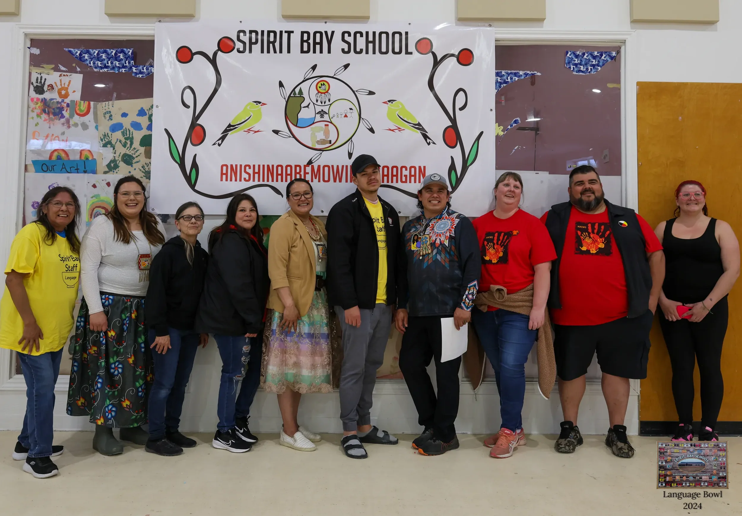 A group of eleven people standing in front of a large banner that reads 'Spirit Bay School' and 'Anishinaabemowin Kaagan'. The group includes individuals in yellow, white, black, beige, and red clothing, some wearing glasses and some smiling. The background features colorful artwork, signs, and a window with blue paper decorations.