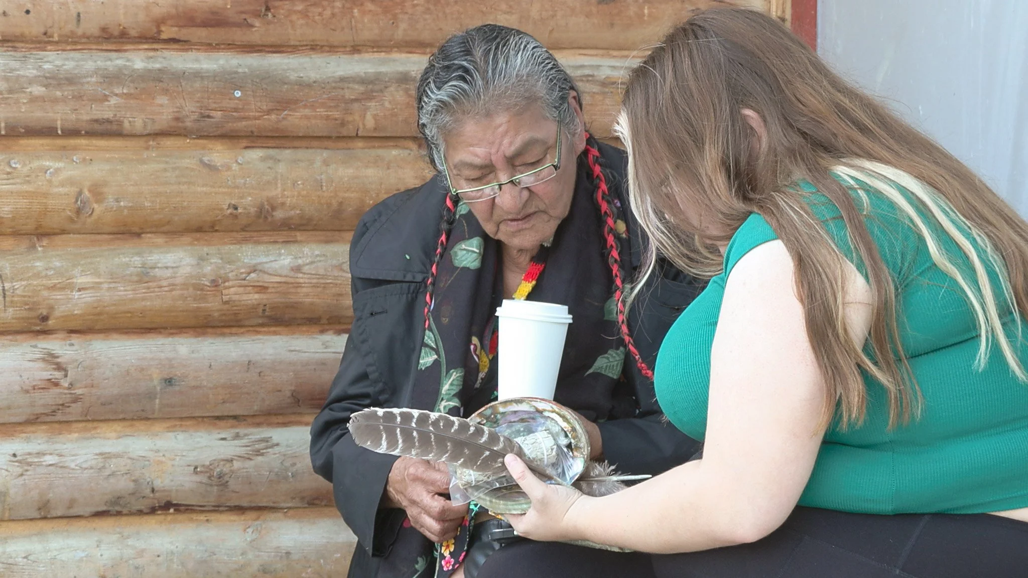 Two women examine a large bird with colorful feathers, one holding it gently while the other observes closely, sitting in front of a wooden log wall.