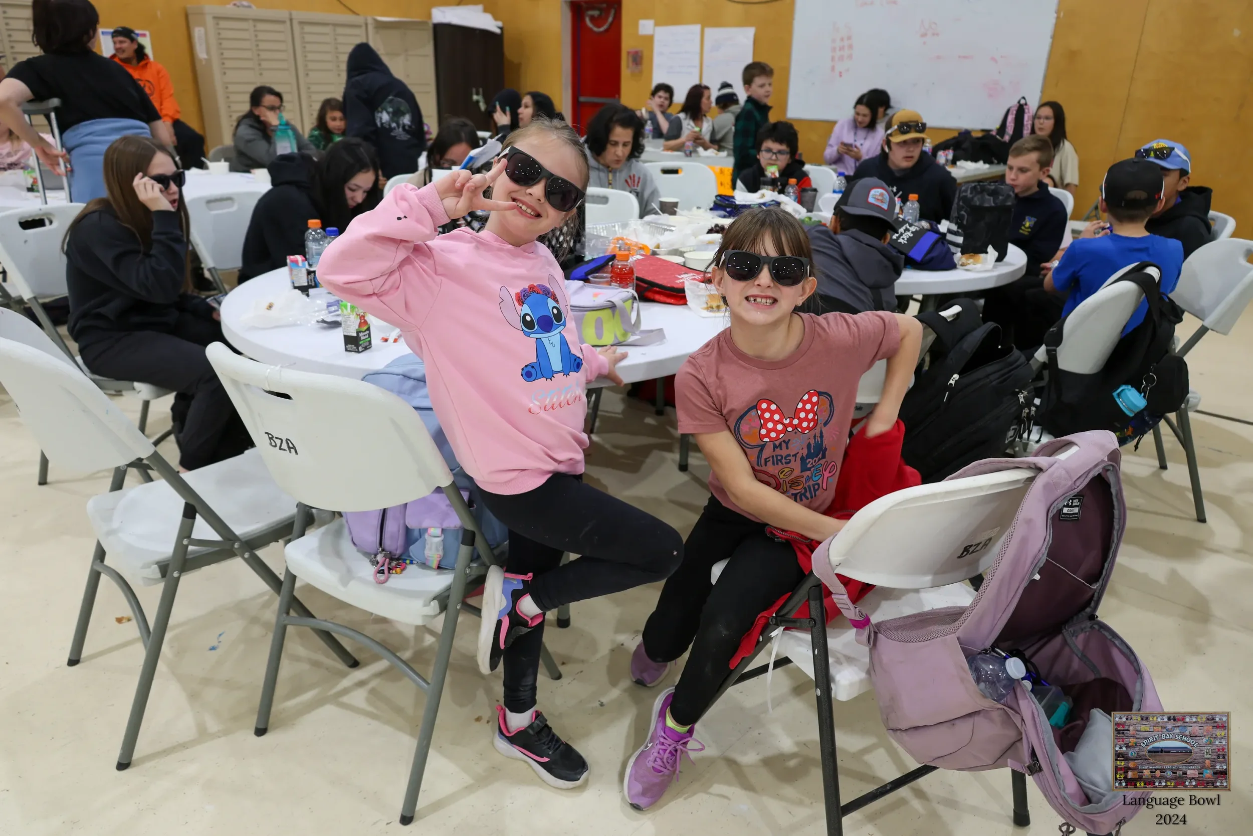 Two young girls wearing sunglasses and smiling, posing for a photo at a large indoor gathering with many children seated at tables in the background.