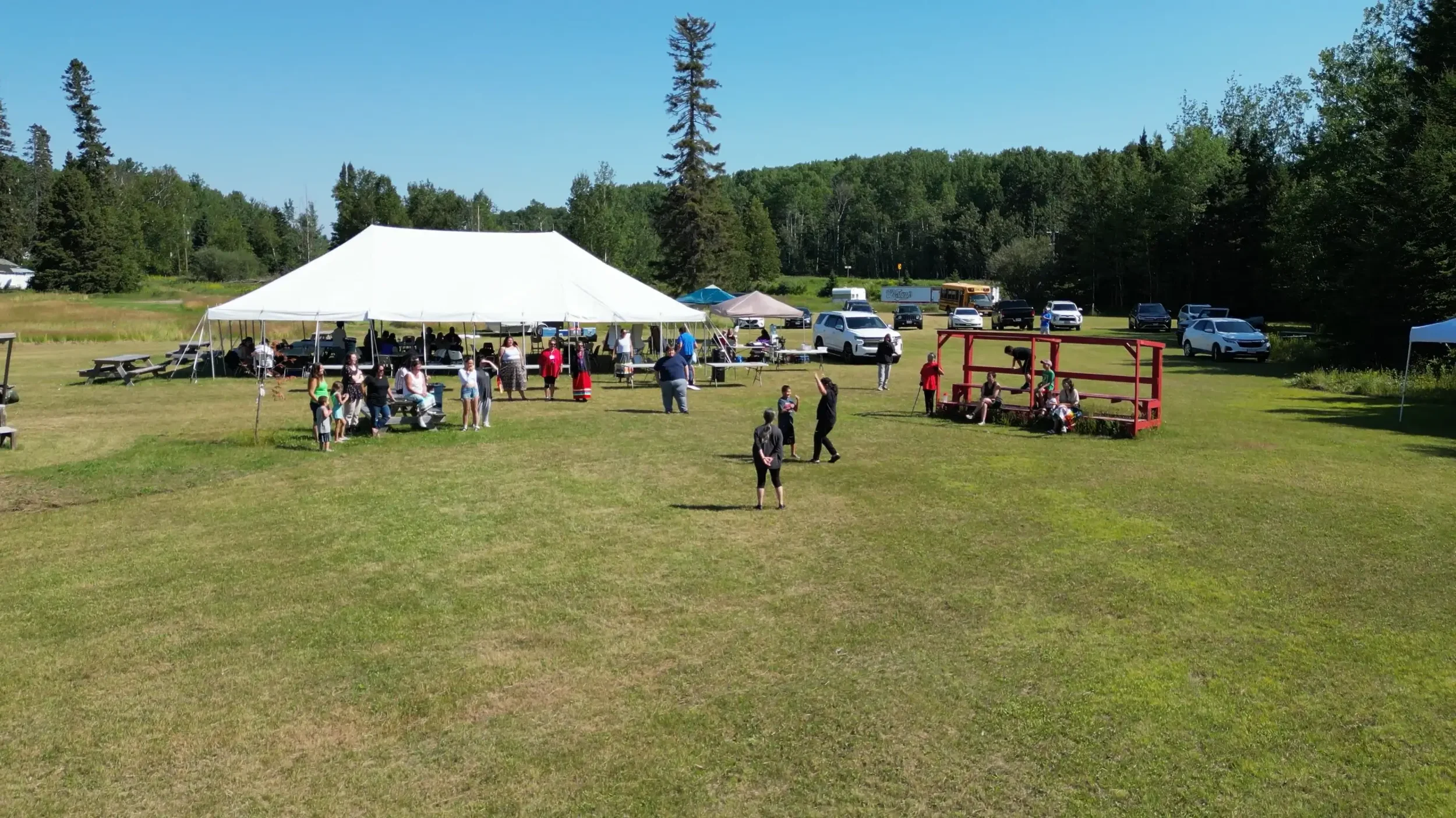 Outdoor gathering with a large white tent, people socializing, and parked cars on a grassy field surrounded by trees on a sunny day.