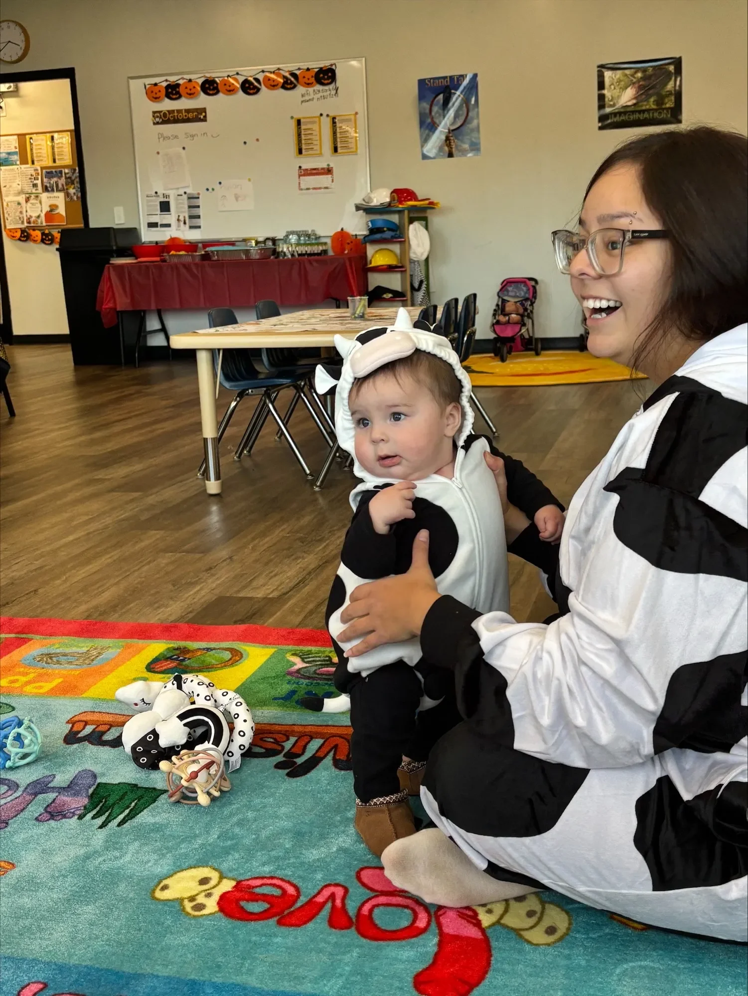 A woman and a baby dressed in cow costumes are sitting on a colorful children's rug in a classroom decorated for Halloween.