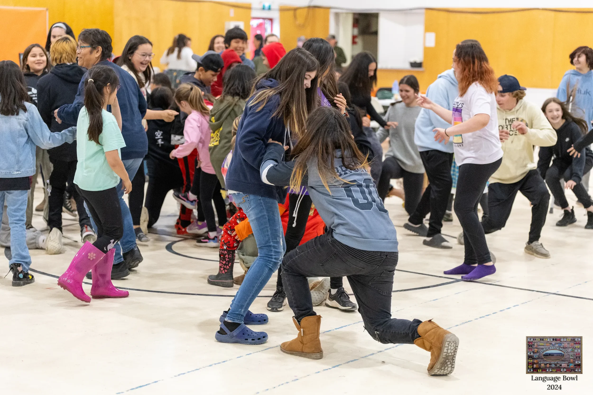 Children and teenagers dance and have fun in a gymnasium during a school event. They are dressed casually, with some wearing rain boots. The background features wooden walls and a yellow accent wall.